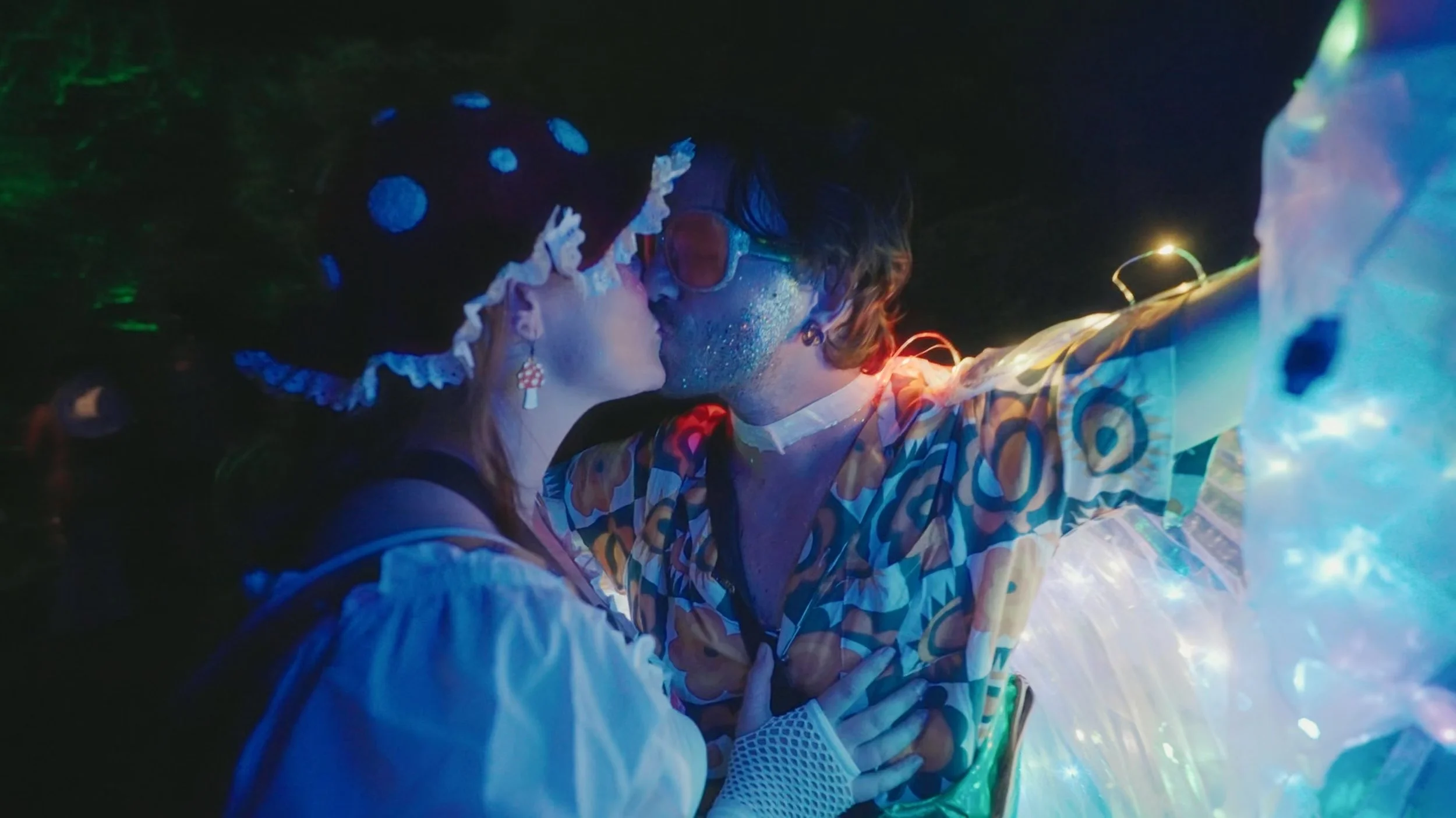 Two people, dressed in colorful, eclectic costumes, are kissing in a dark setting illuminated by vibrant, multicolored lights, creating a surreal, festive atmosphere.