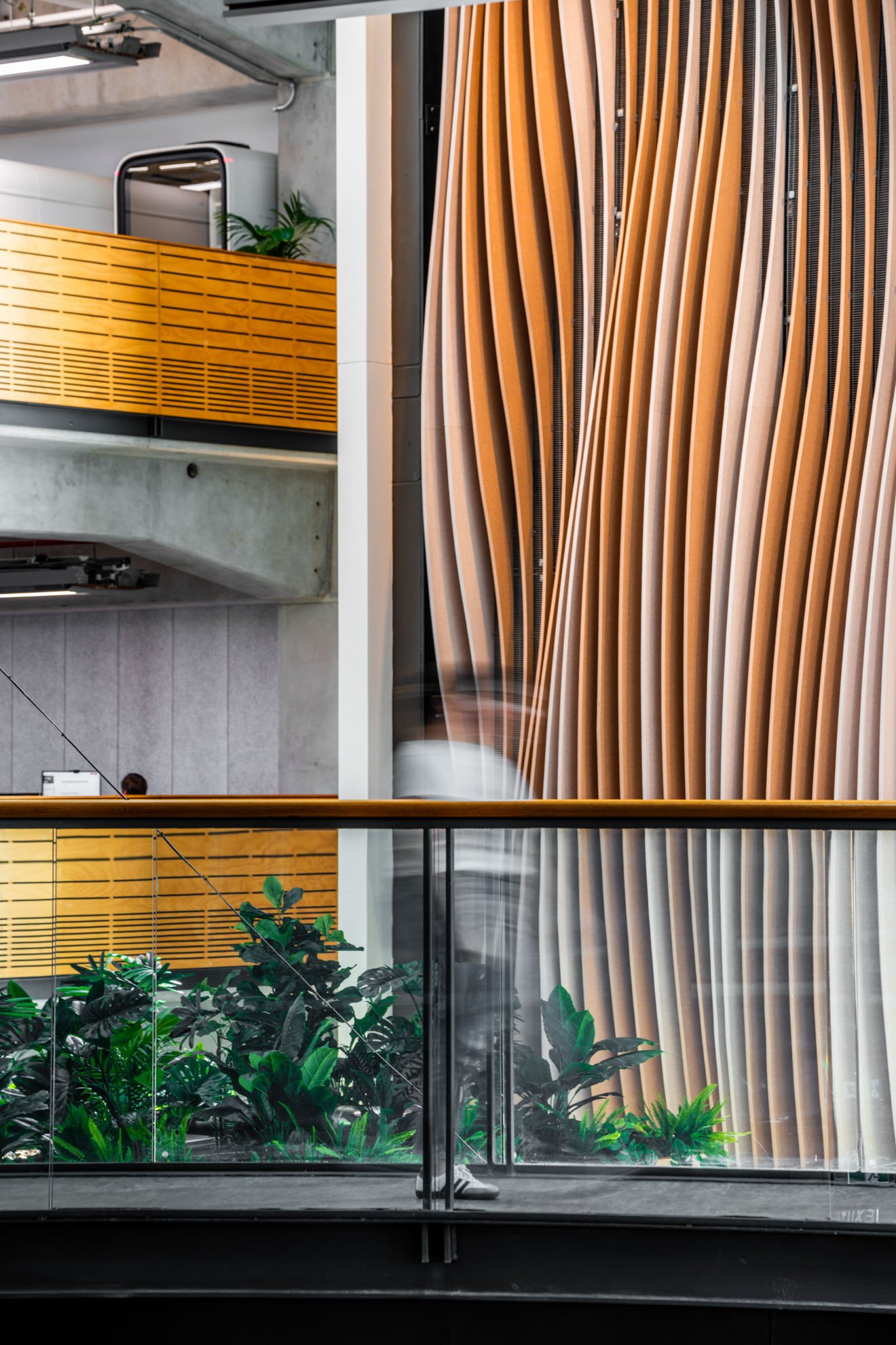 Interior of a modern building with decorative wooden wall panels, glass railing, and lush green plants on a lower level.