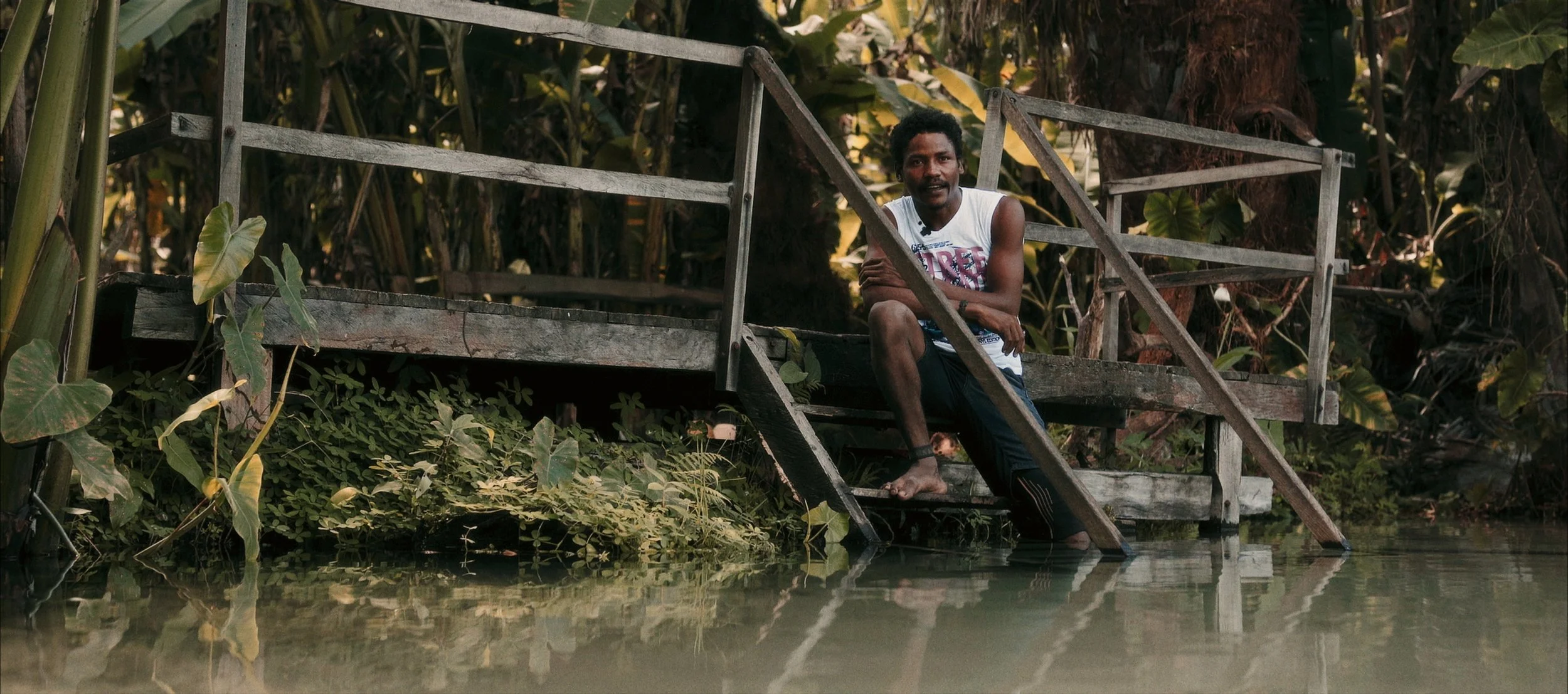 A man sitting on a wooden dock surrounded by lush green plants and trees, with water reflecting the scene.