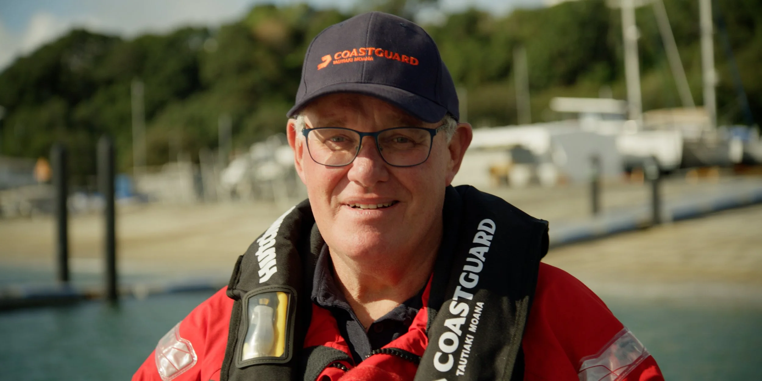 A man in Coast Guard gear smiling outdoors near a marina with boats in the background.