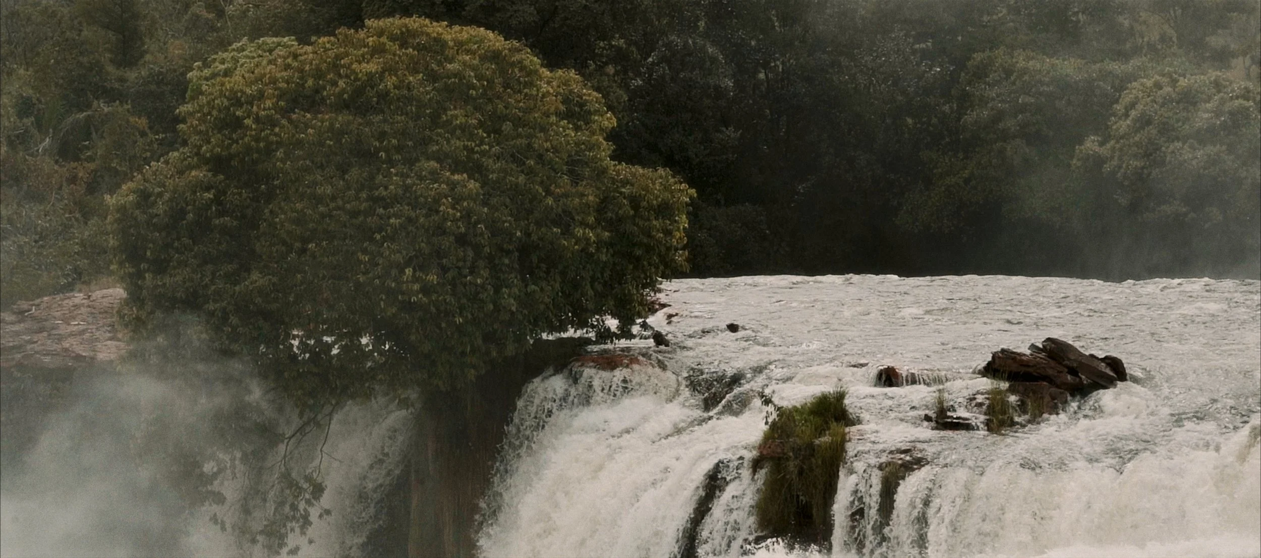 A waterfall flowing over rocks with lush green trees and vegetation in the background, mist rising from the cascading water.
