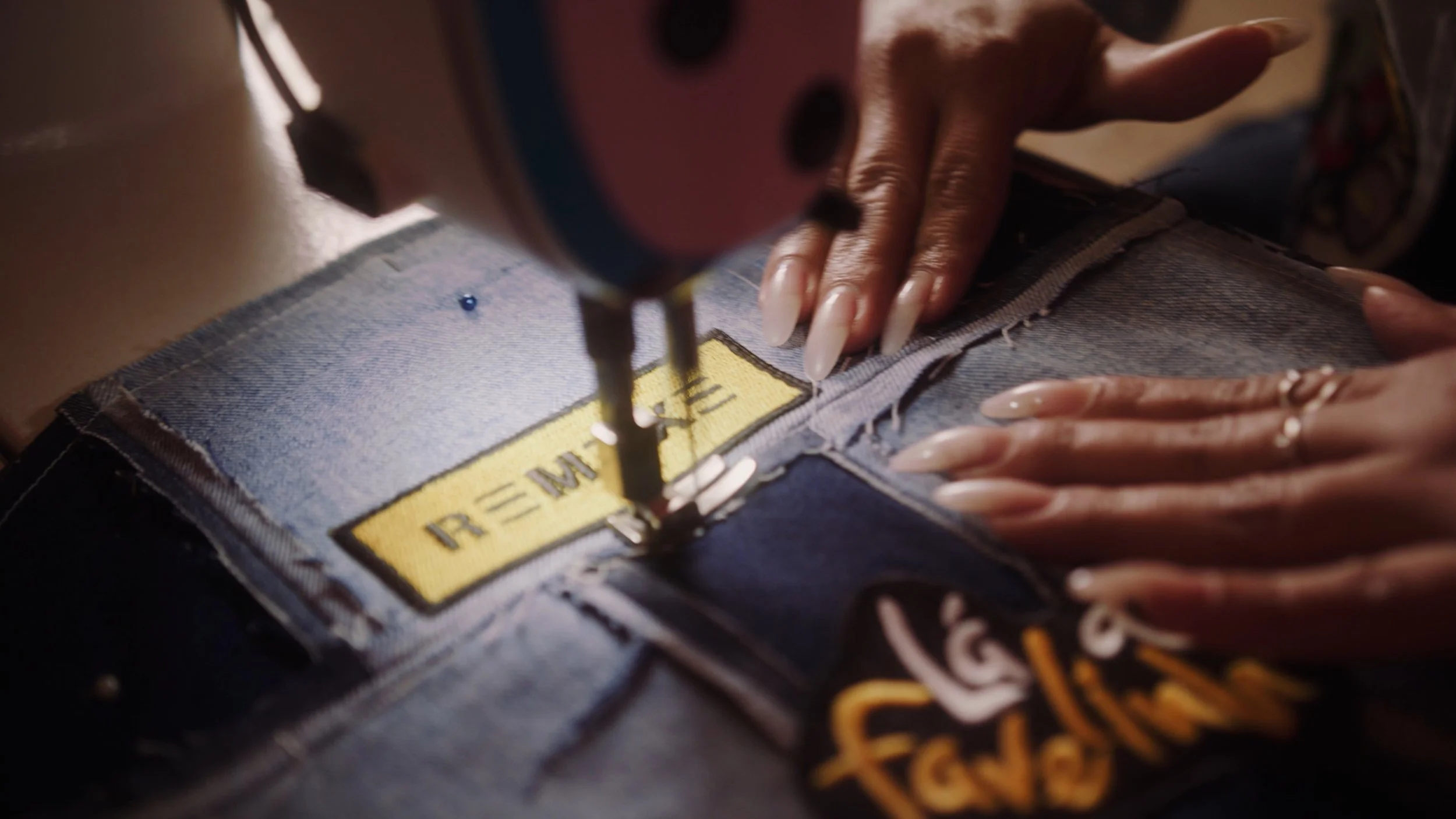 Close-up of a person's hands sewing a yellow patch with the word 'RE-MAKE' onto denim fabric using a sewing machine.