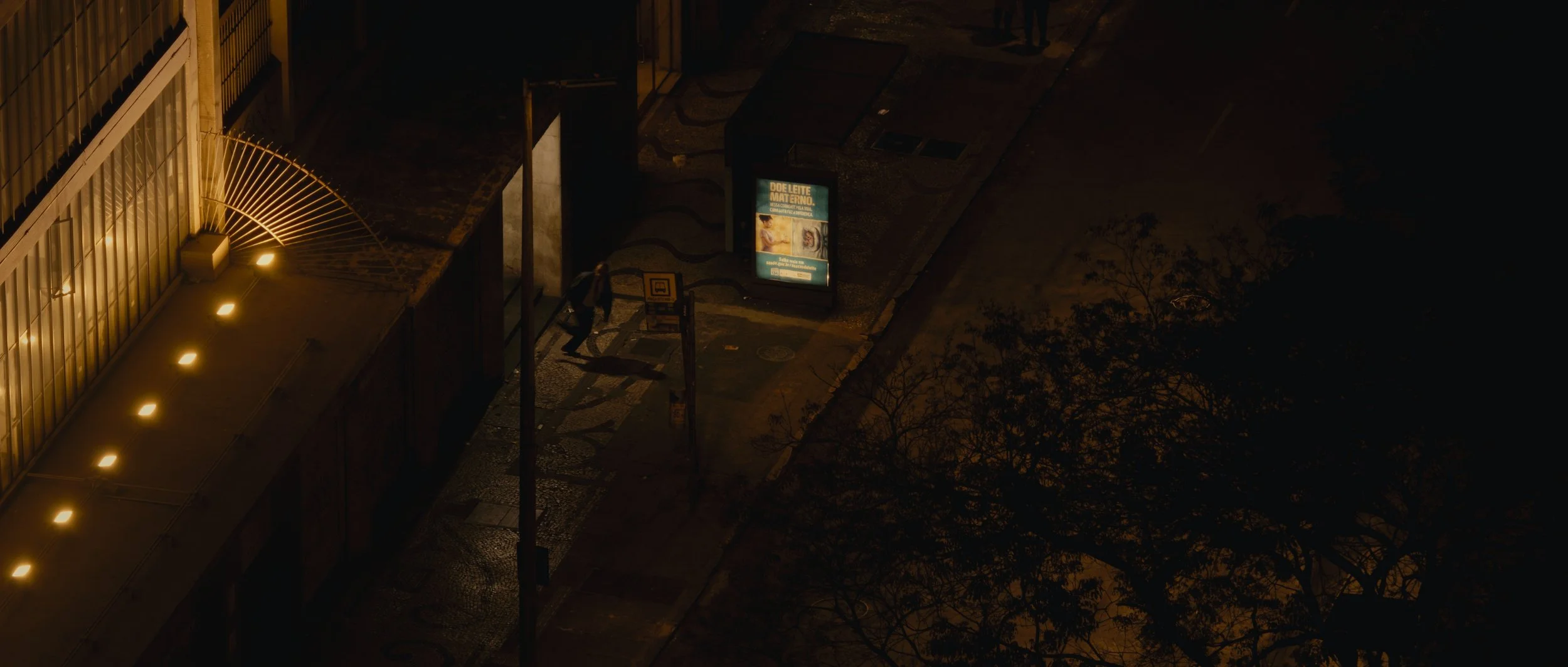 A city street at night with a person walking past a lit advertisement display and streetlights, surrounded by buildings and trees.