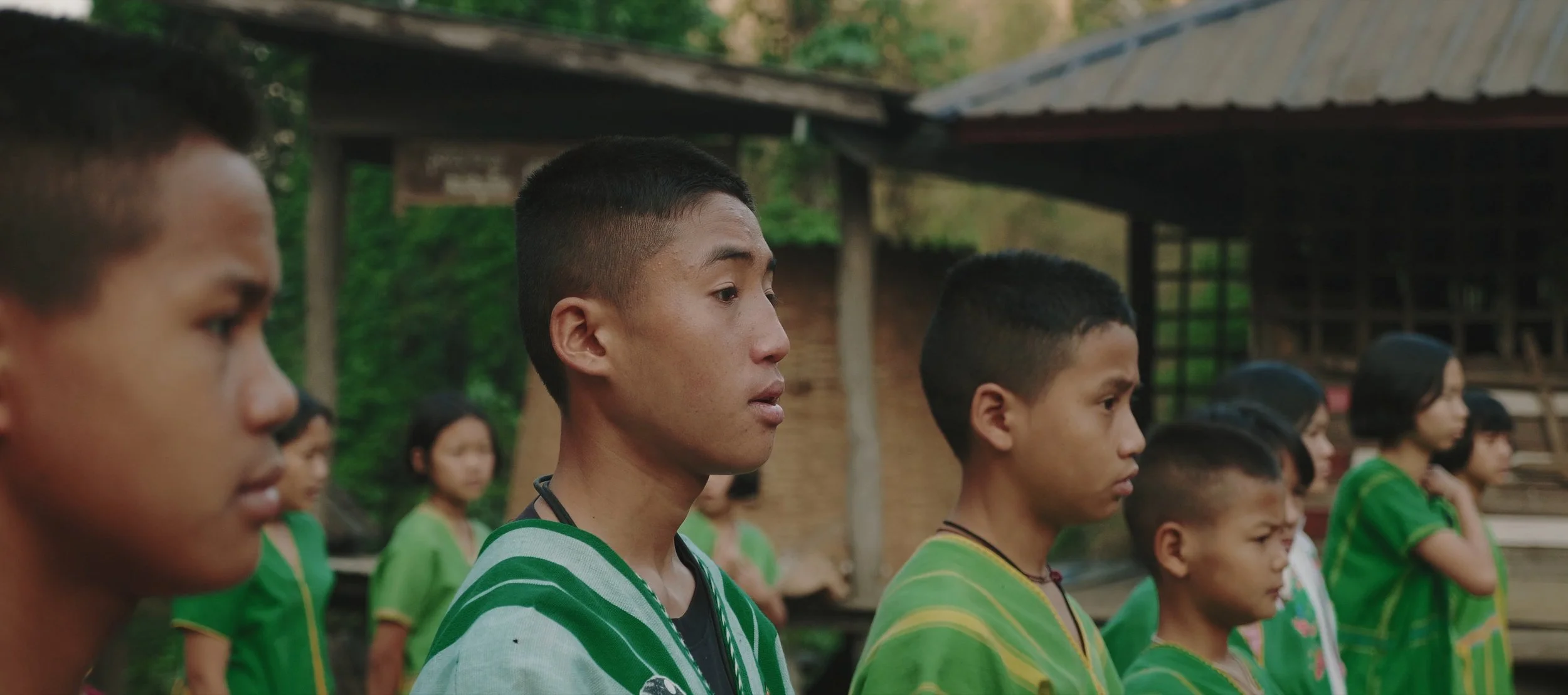 A group of young people wearing green shirts standing outdoors with a wooden structure and trees in the background.