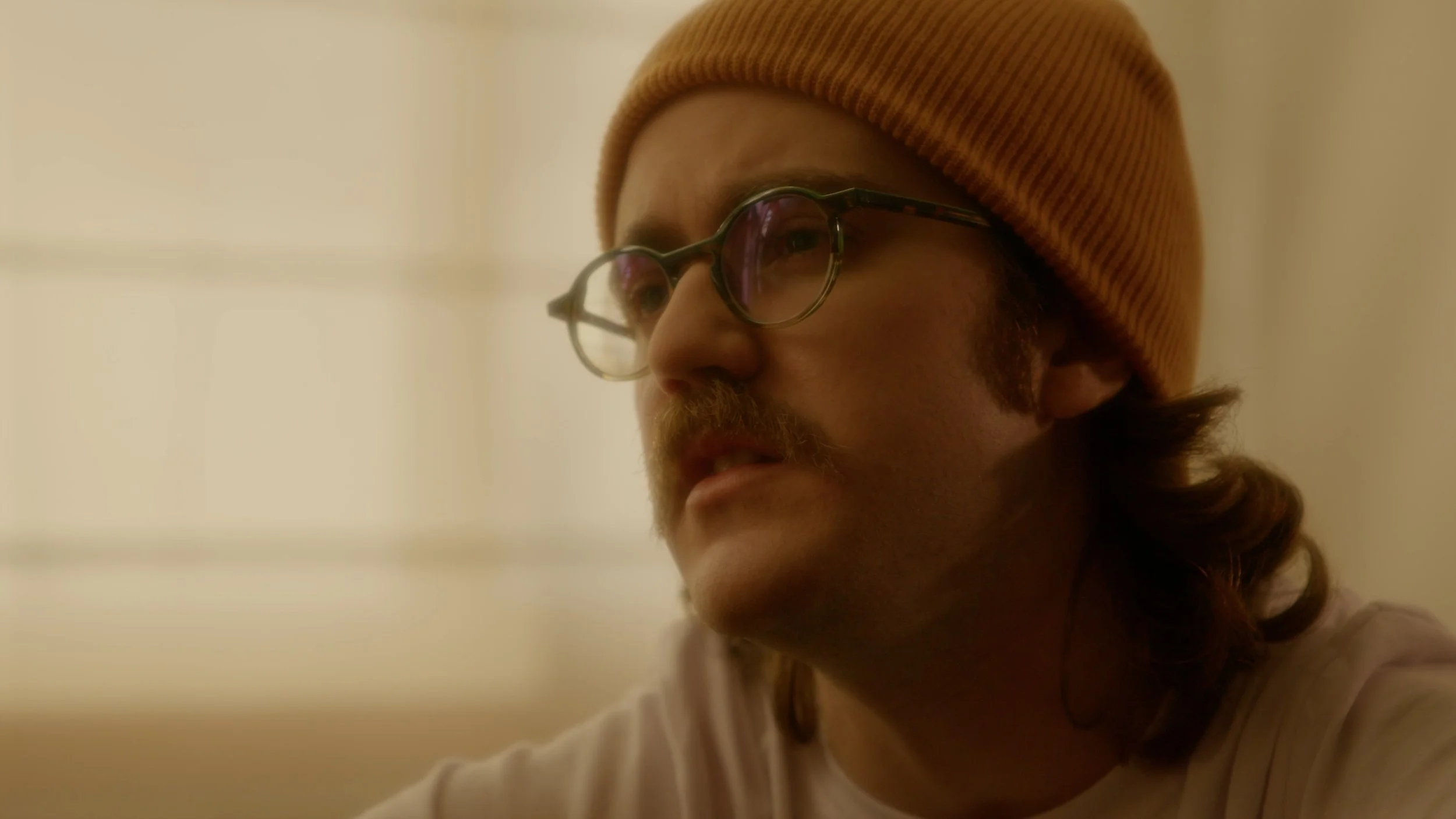 Close-up of a man wearing glasses and a tan beanie, with shoulder-length brown hair and a mustache, looking pensively to the side.