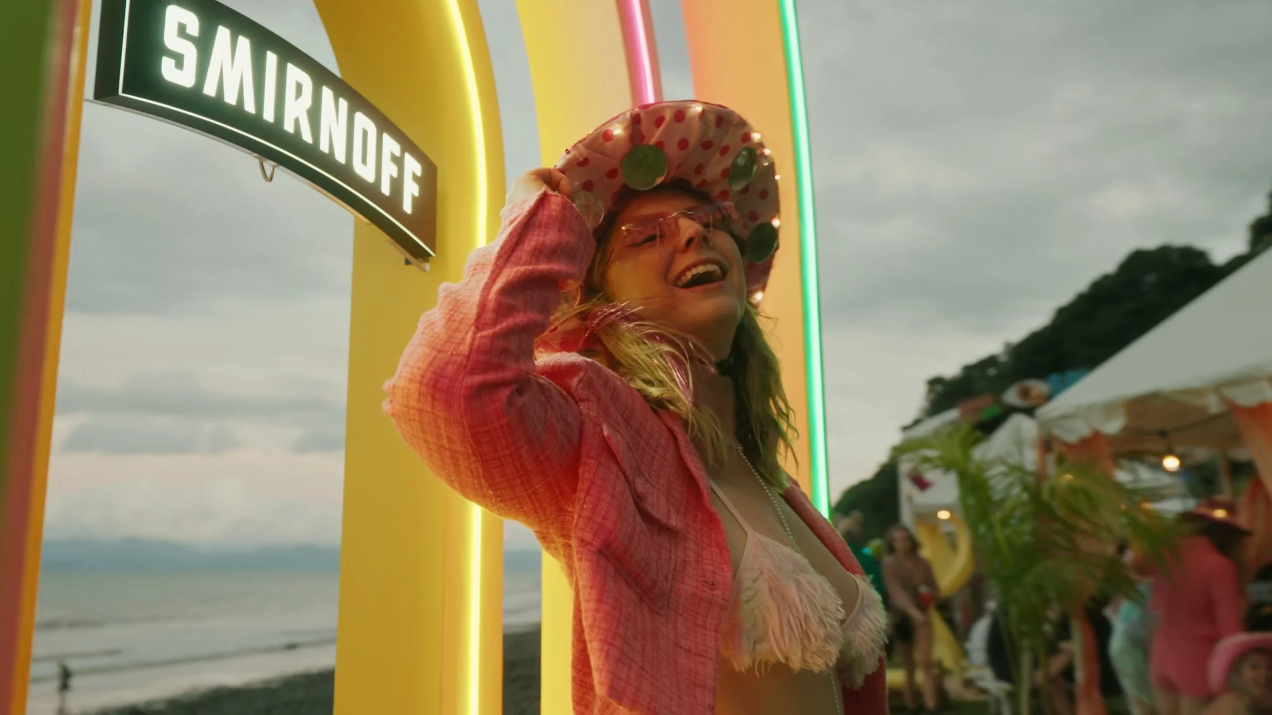 A young woman at a beachside amusement park, smiling, wearing a polka dot hat decorated with green circles, pink glasses, a pink jacket, and a feathered top. There are tents and people in the background, with a wet beach and cloudy sky.