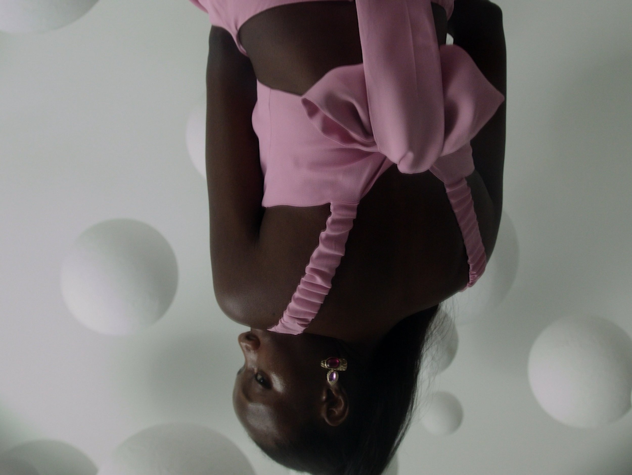 A woman wearing a pink outfit with ruffles, earrings, and dark skin, in front of a white background with white spherical decorations.