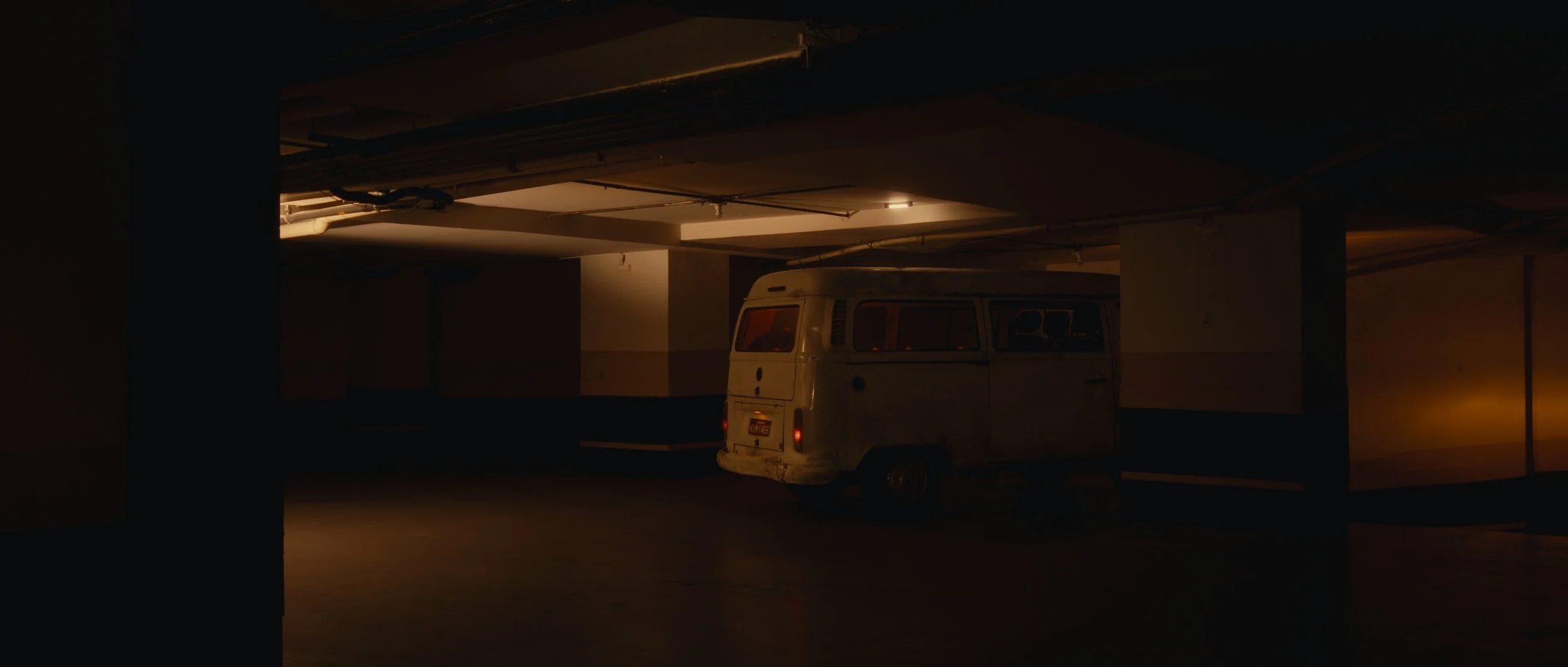 An old, white van parked in a dimly lit underground parking garage with minimal lighting and dark shadows.