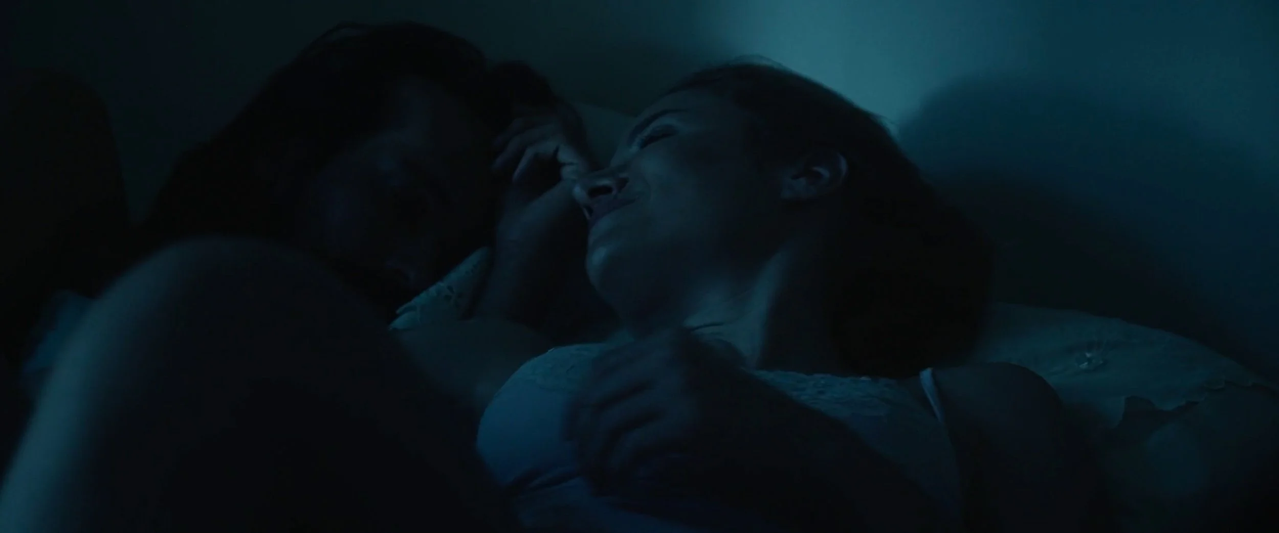 A man and a woman lying close together in bed, sharing an intimate moment in a dimly lit room.