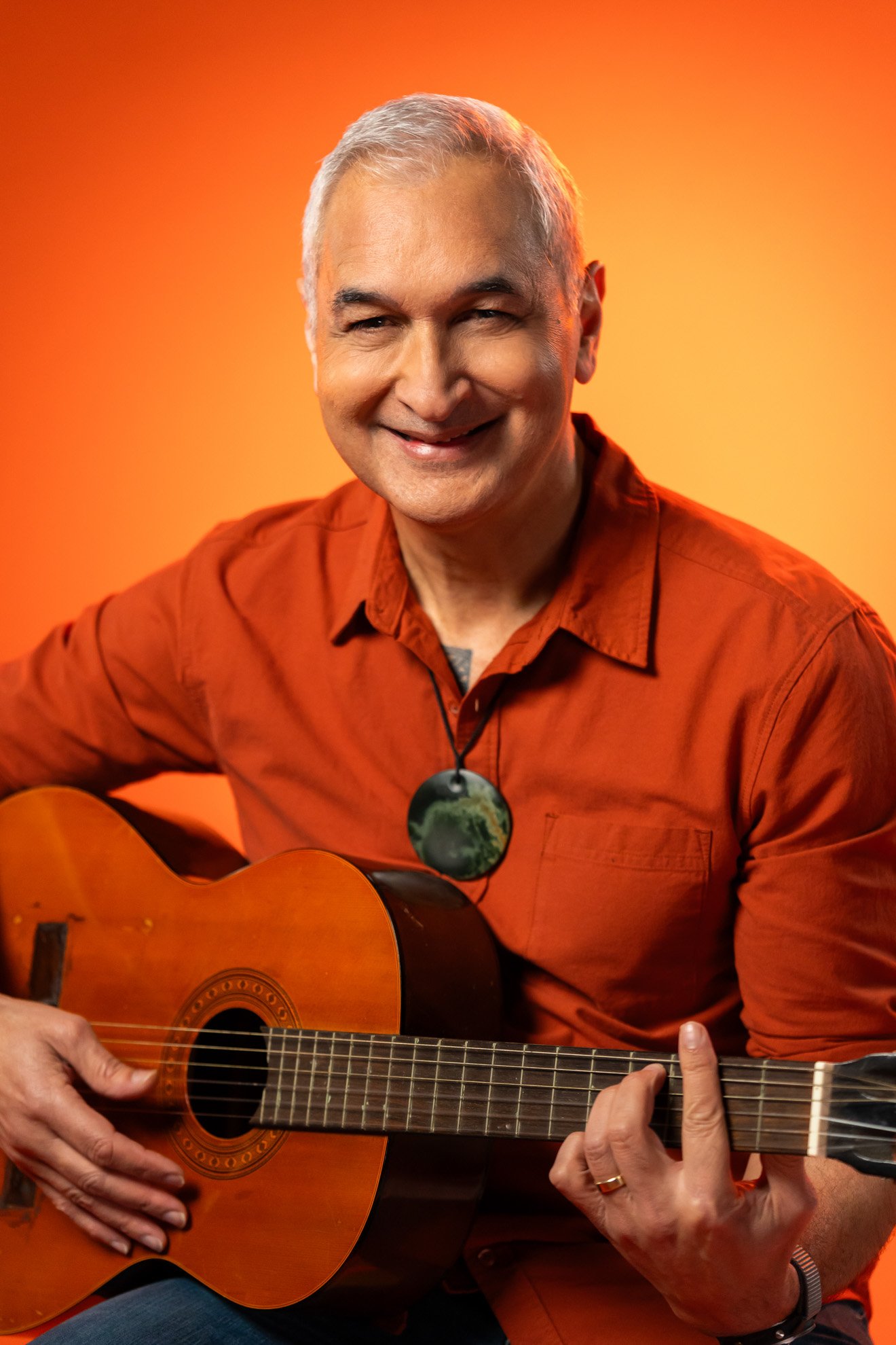A smiling middle-aged man with gray hair wearing an orange shirt playing an acoustic guitar against an orange background.