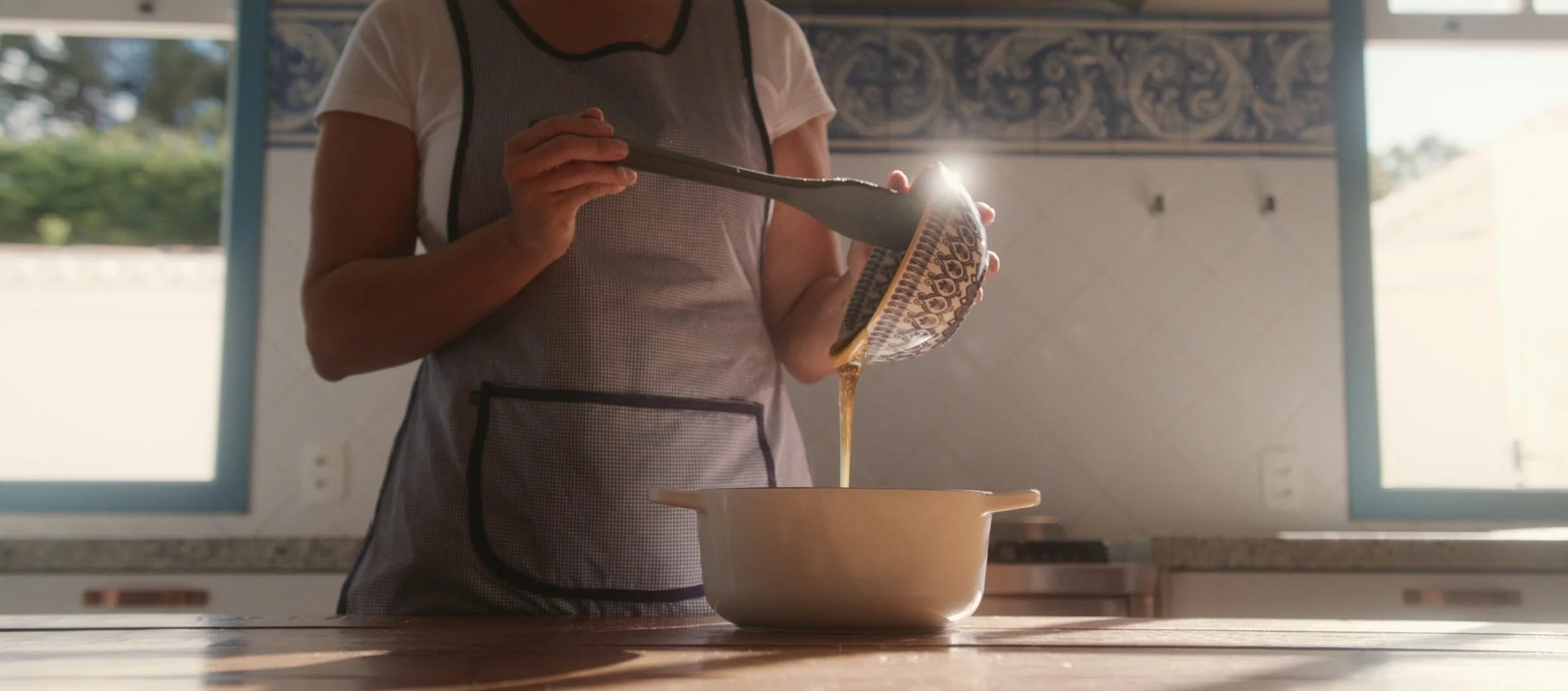 A person pouring liquid from a patterned bowl into a beige baking dish in a bright kitchen.