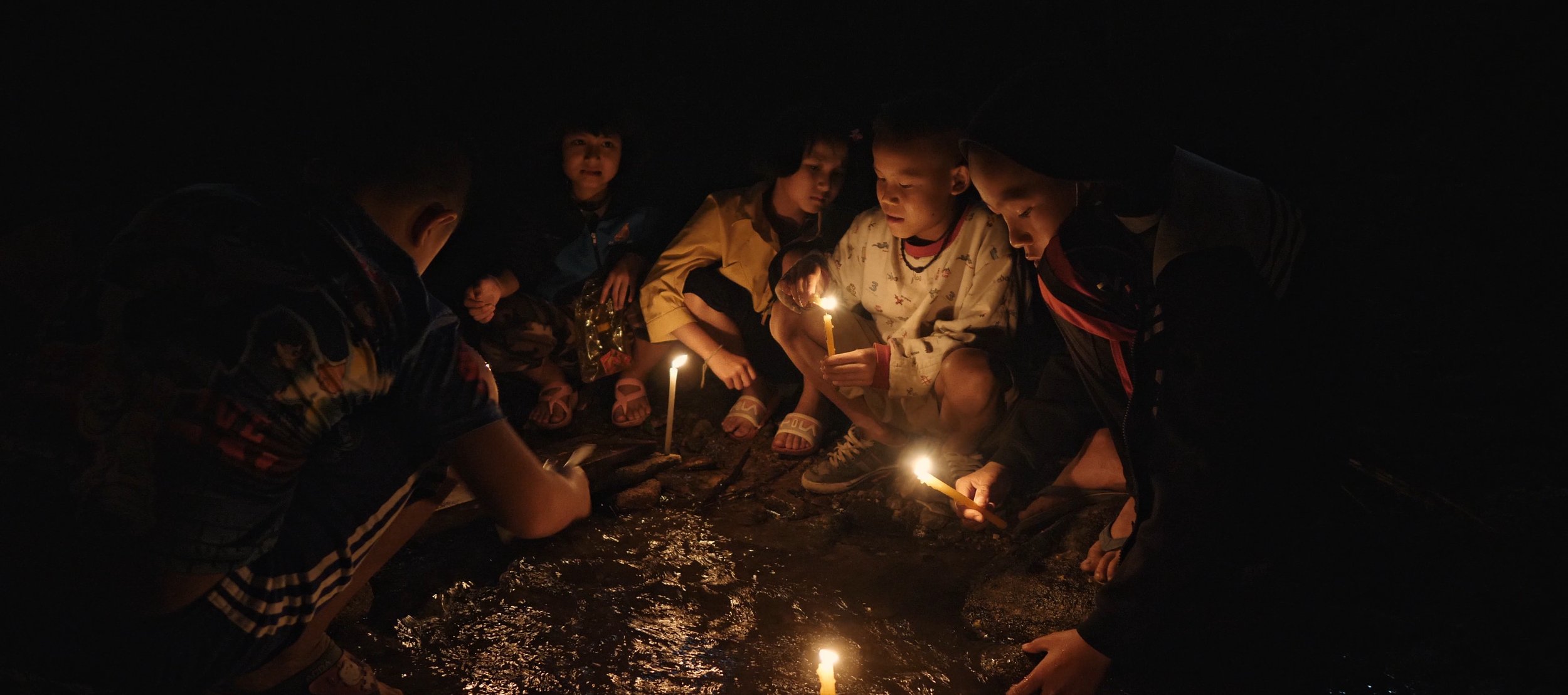 Group of children gathered around a small fire outdoors at night, holding candles.