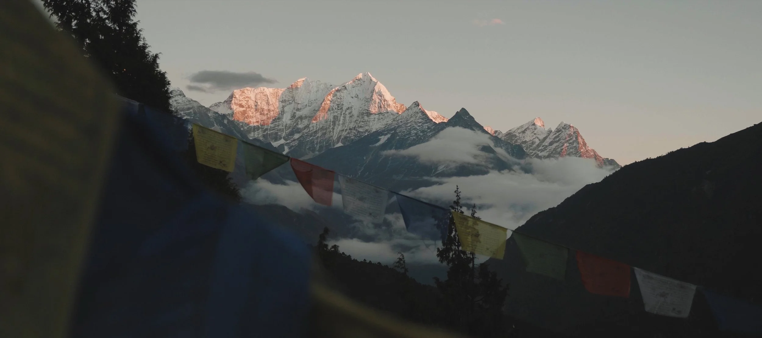 Mountains with snow-capped peaks at sunrise or sunset, with prayer flags in the foreground.