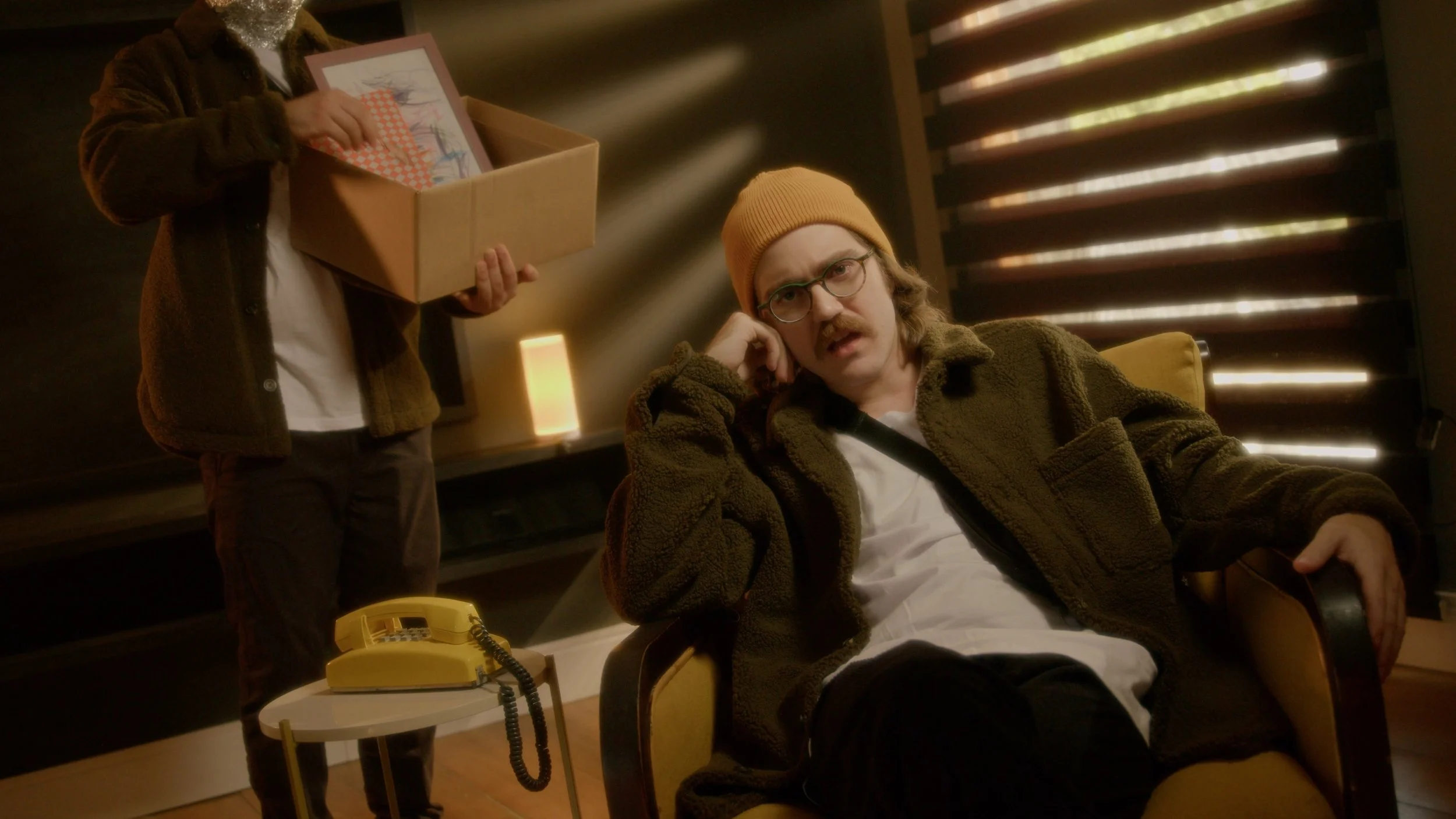 A man with glasses, a mustache, and wearing a yellow beanie and brown jacket sits in a yellow armchair, looking confused or bored. Behind him, another person stands, holding a cardboard box full of framed pictures. A yellow rotary phone is on a small