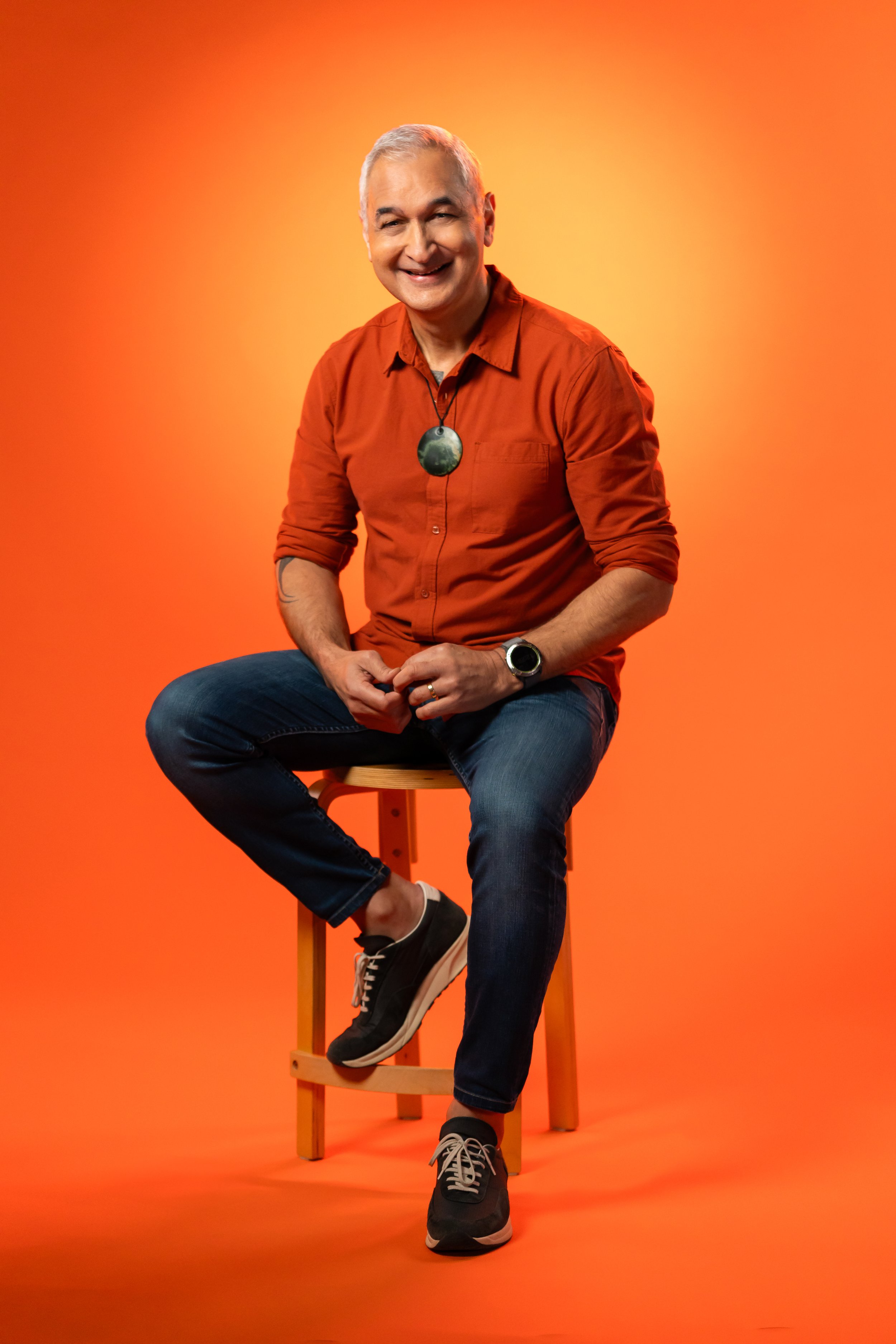 Smiling middle-aged man with gray hair wearing a burnt orange shirt, dark jeans, and black sneakers, sitting on a wooden stool against a orange background.