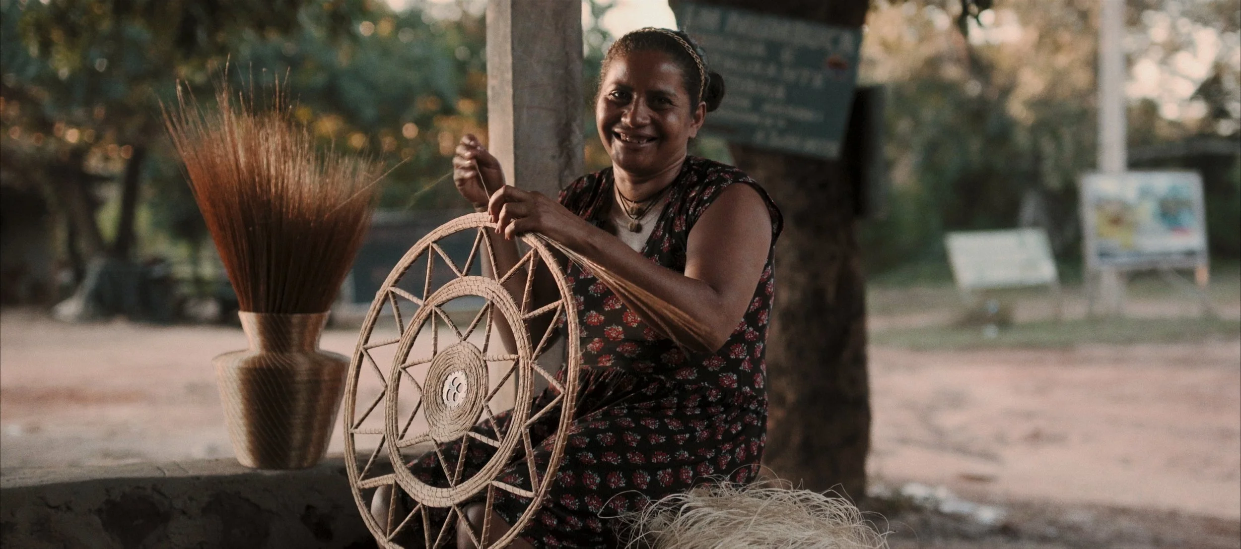 A woman sitting outdoors, weaving a dreamcatcher with a smile, surrounded by traditional craft materials and a large vase of dried grass.