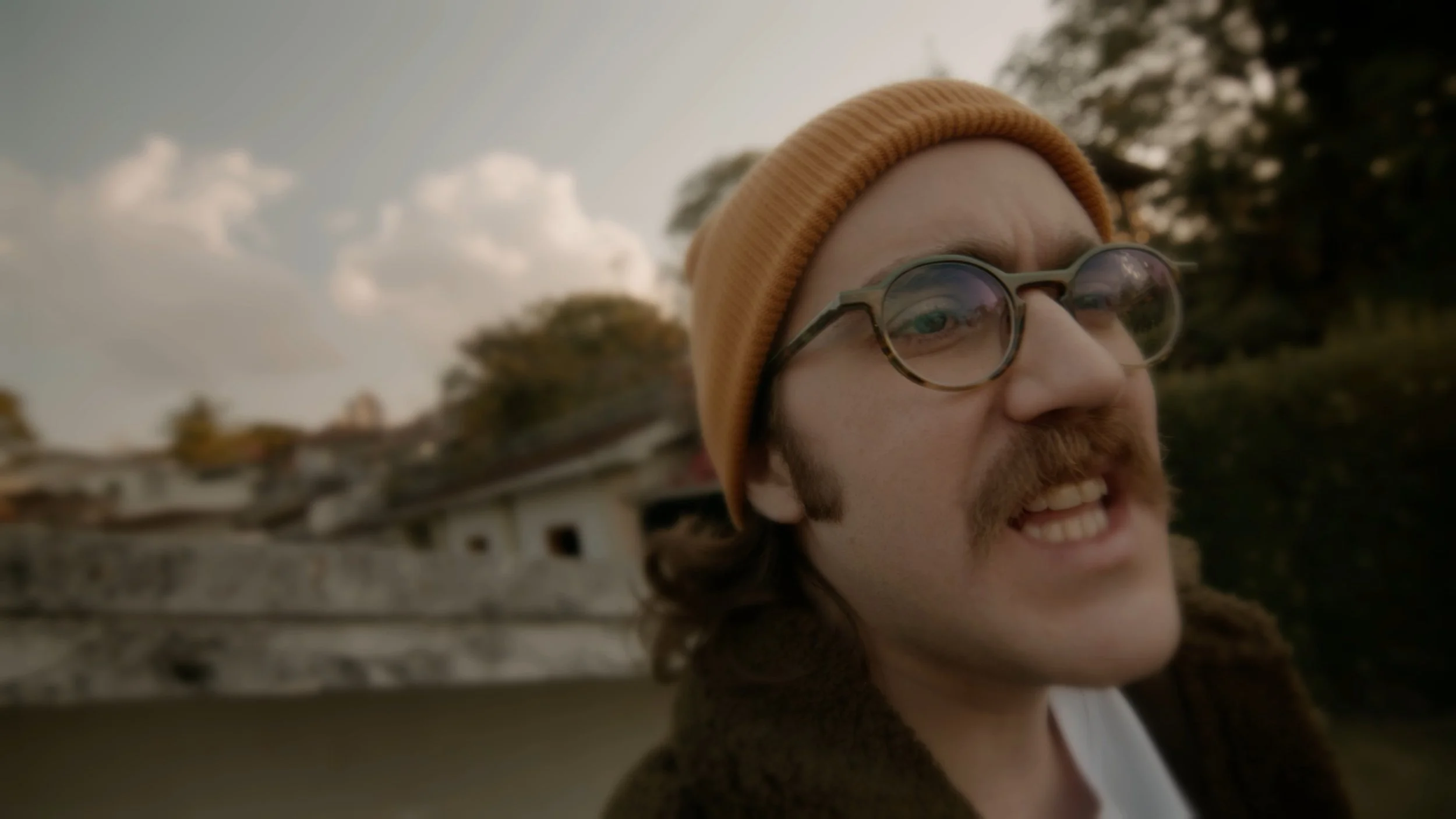 A young man with glasses, a mustache, and a beard wearing an orange beanie, smiling, outdoors with a cloudy sky and trees in the background.