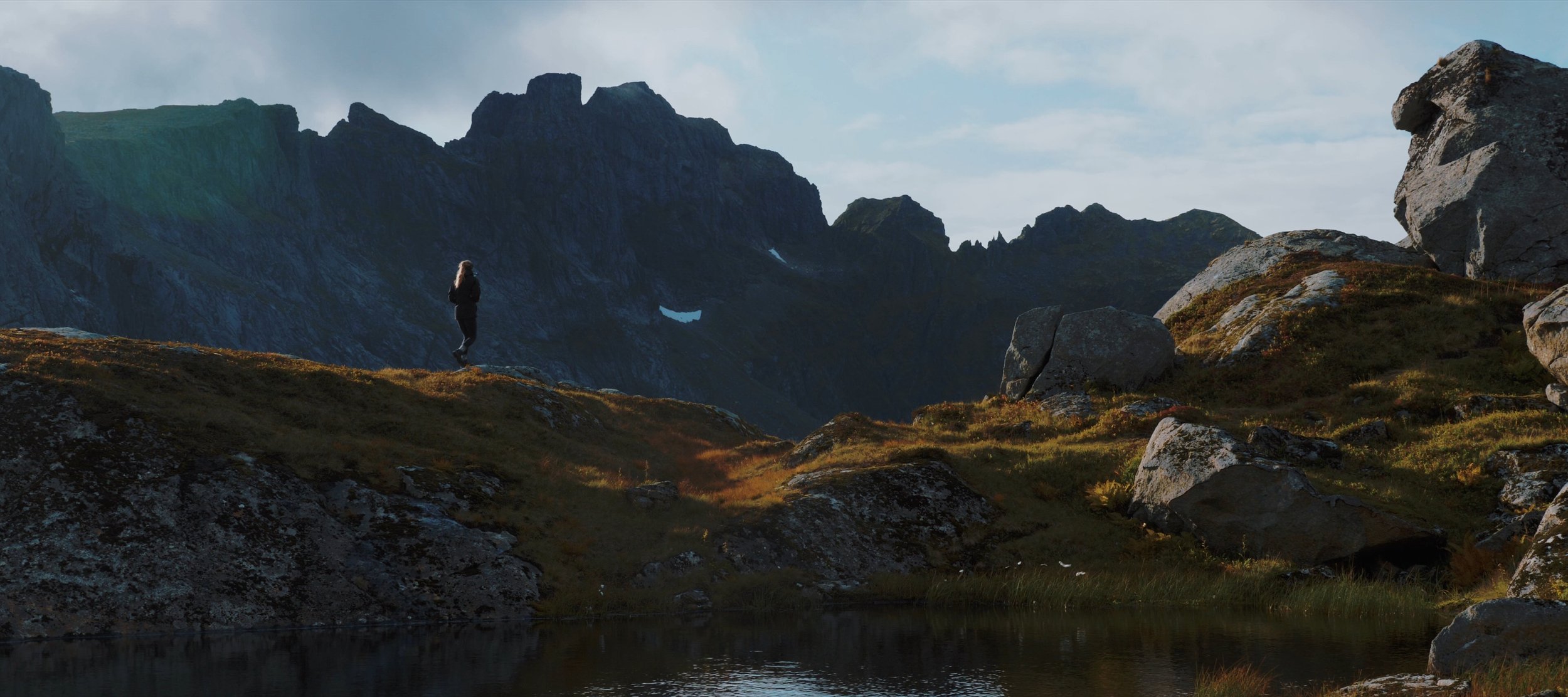 A person standing on a grassy hill overlooking rocky mountains and a small body of water, with a cloudy sky overhead.