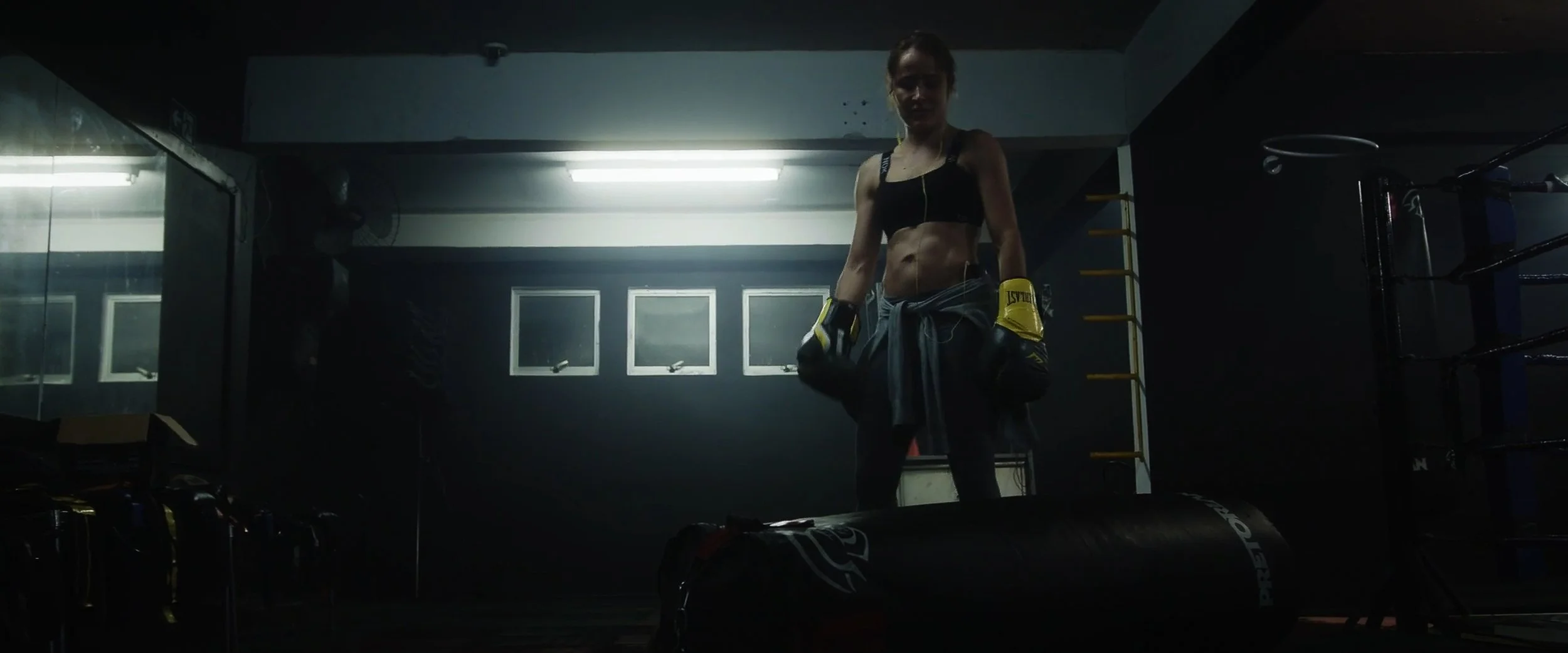A female boxer in a gym, wearing boxing gloves and workout clothes, standing next to a punching bag, with gym equipment in the background.