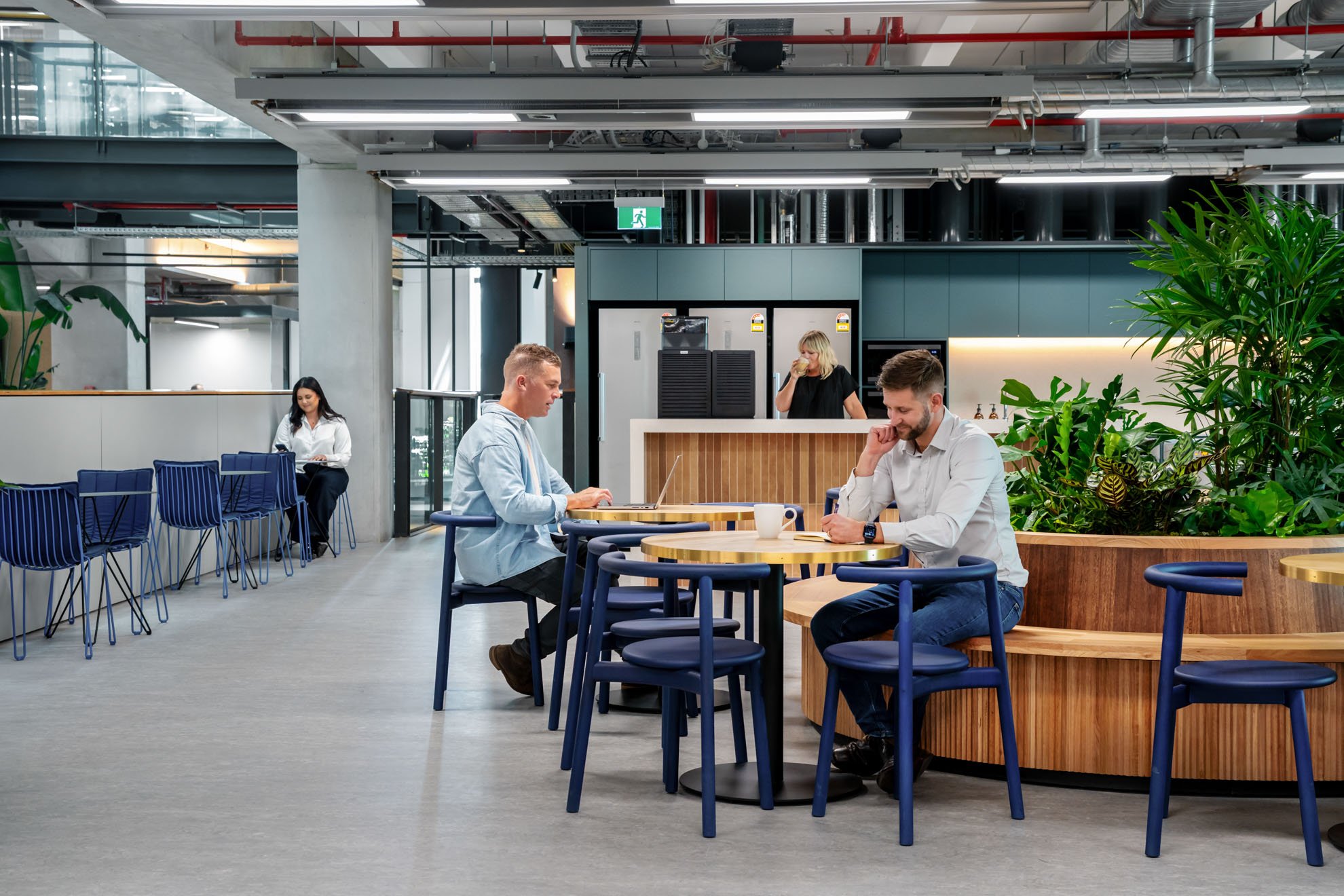 People sitting at tables in a modern, open-plan cafe or coworking space with green plants and industrial ceiling design