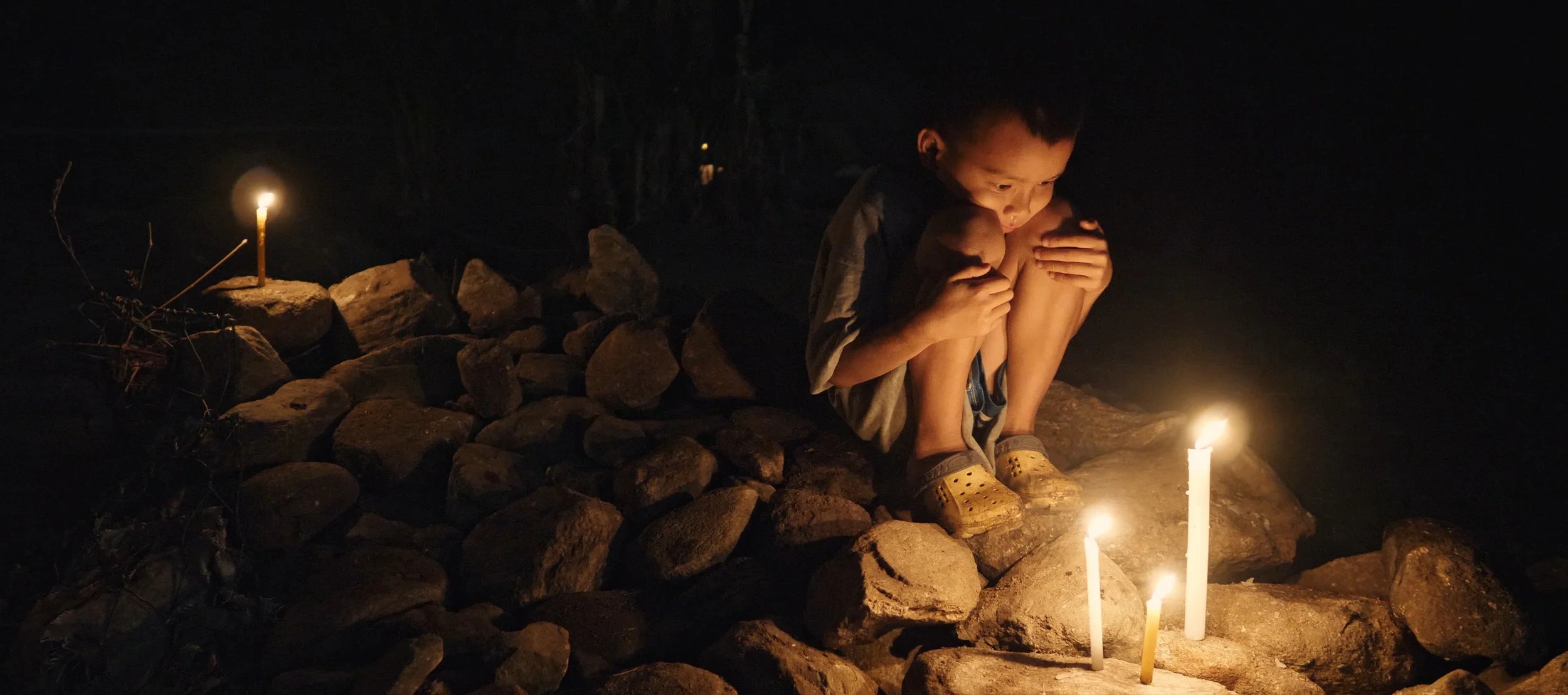 A young boy crouches on a rocky surface at night, illuminated by the light of three candles placed among the rocks, with darkness surrounding him.