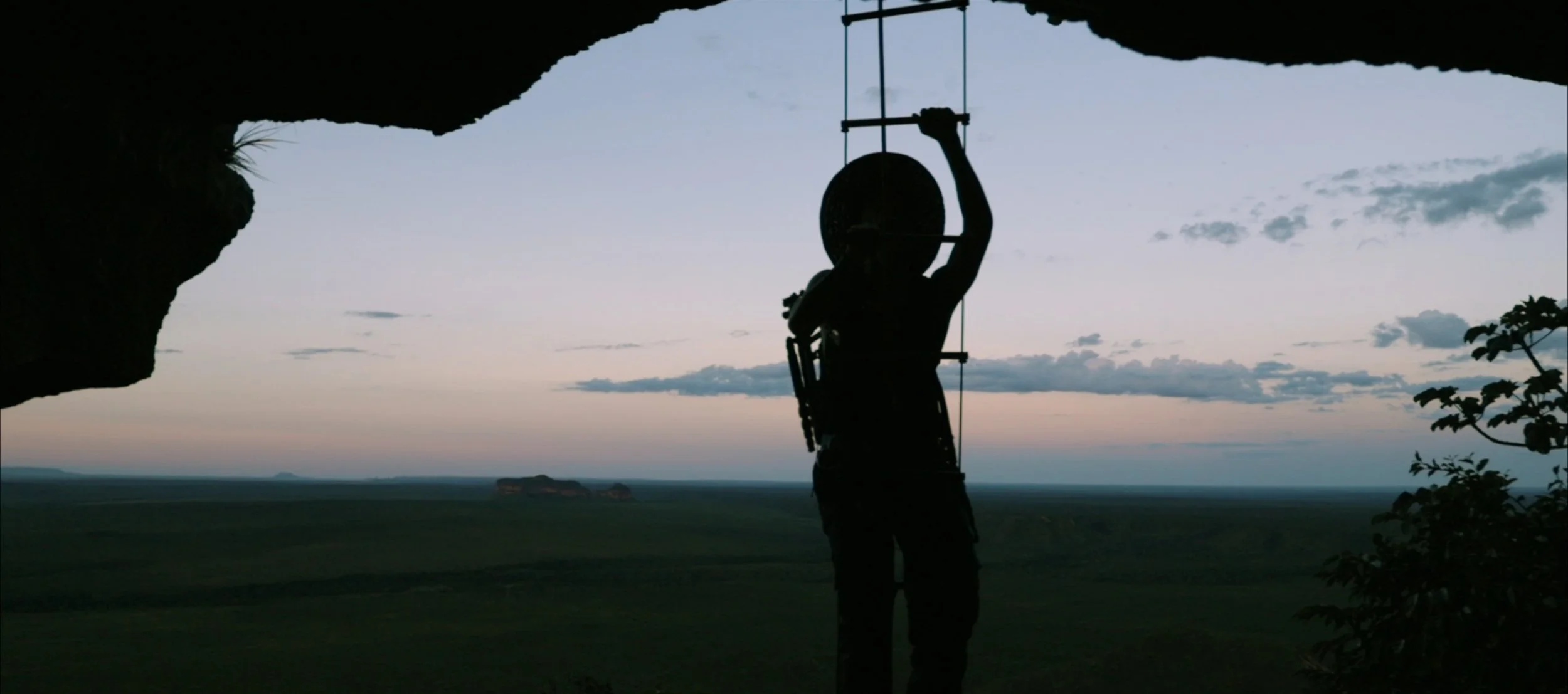 Silhouette of a person with climbing gear, standing on a ledge with a view of a vast landscape and a setting sun in the background.
