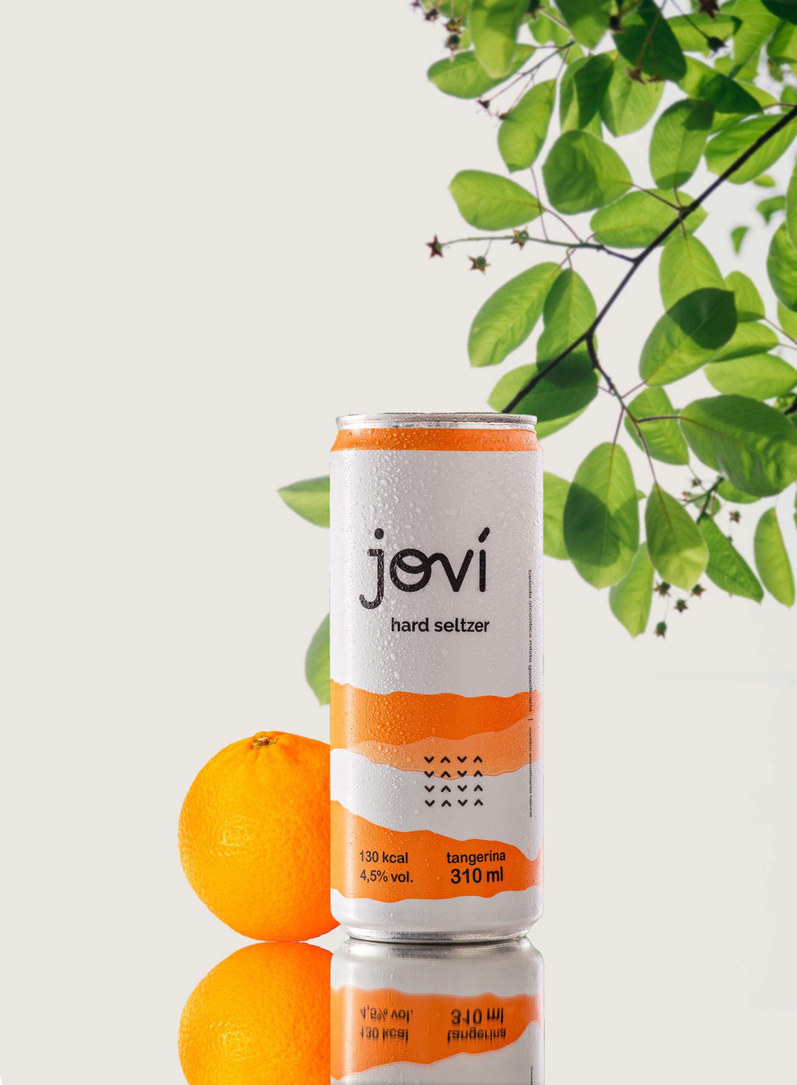 A can of Jovi hard seltzer with orange flavor, two oranges, and a leafy branch in the background, with a reflection of the can and oranges on the surface.