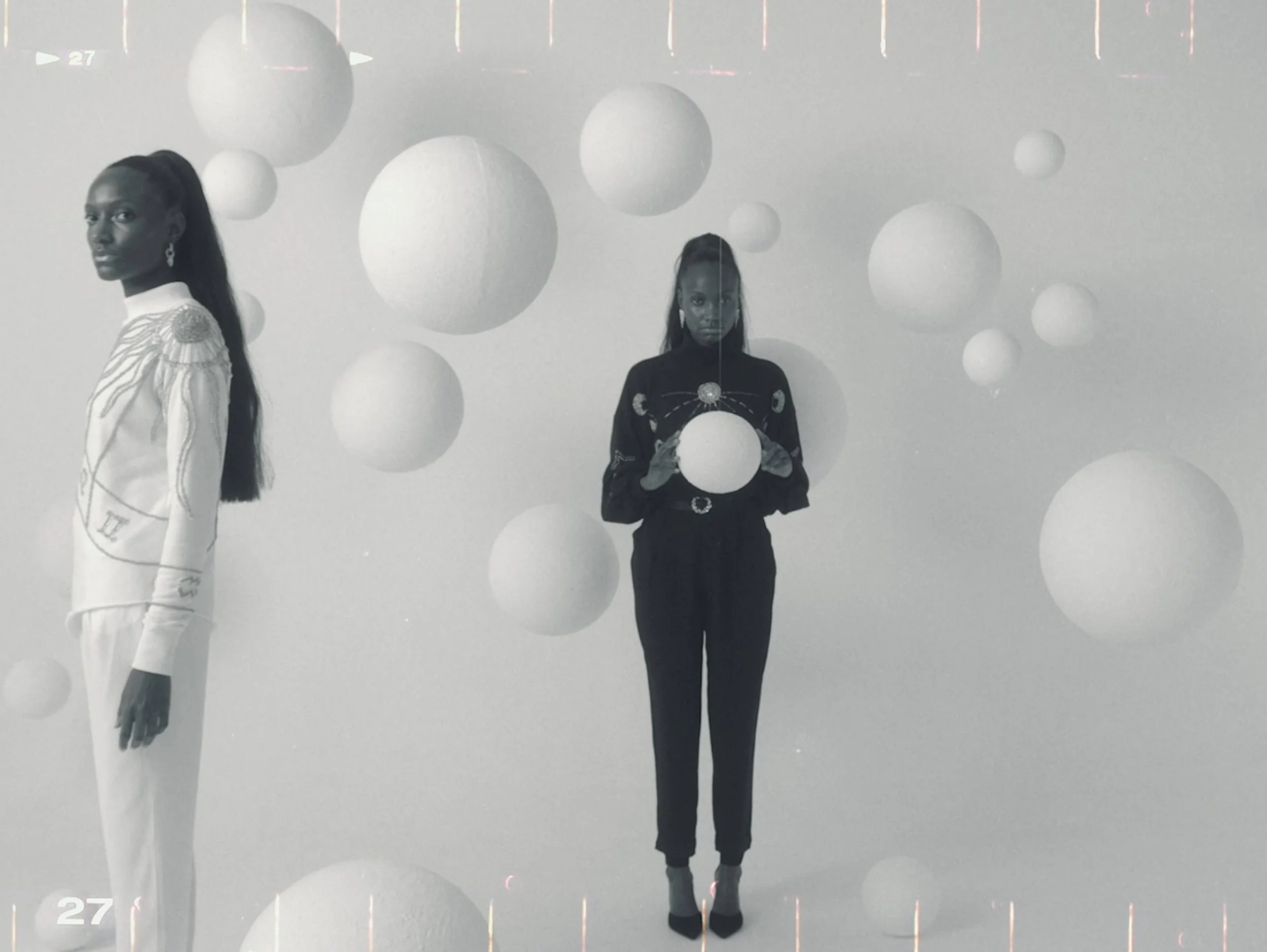 Two women standing among hanging spherical objects in a minimalistic setting, with one woman on the left in light-colored attire and the other on the right in dark clothing holding a sphere.