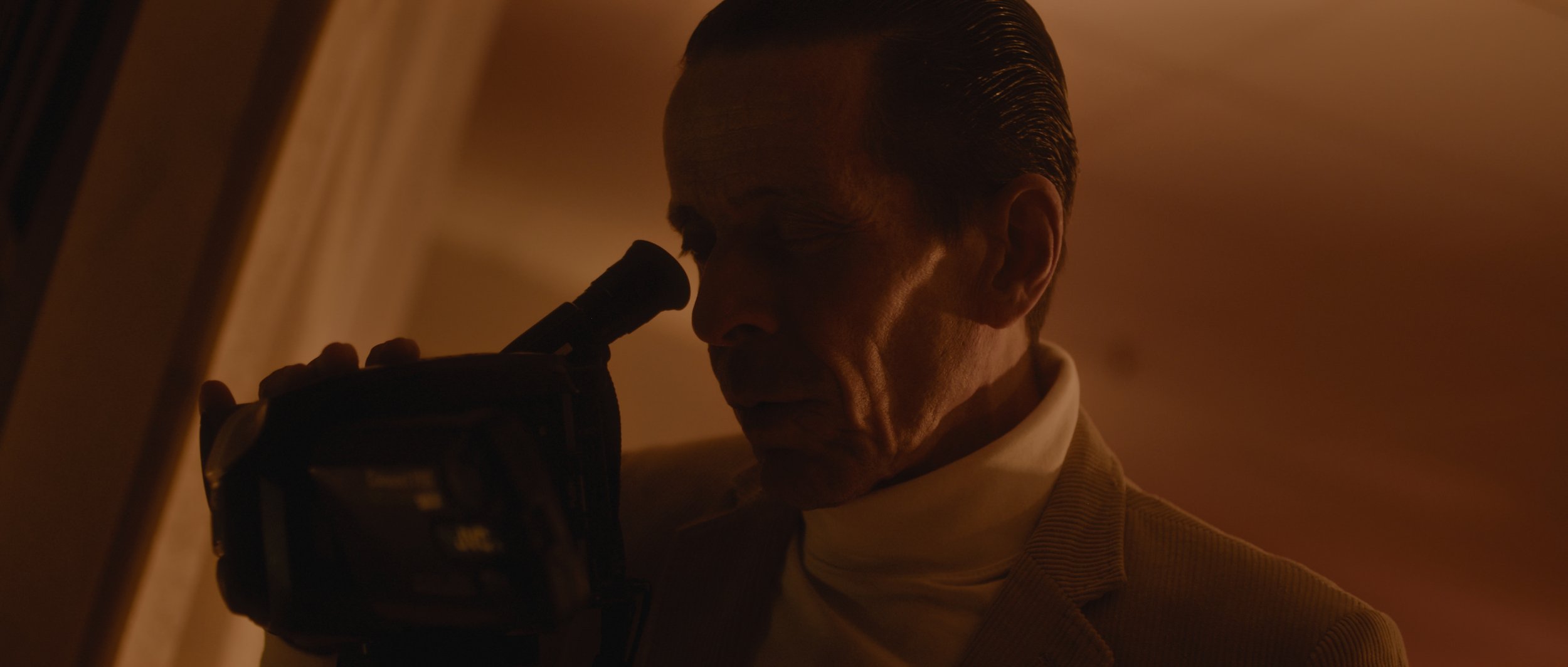 A man with slicked-back hair looking through a microscope in a dimly lit room.