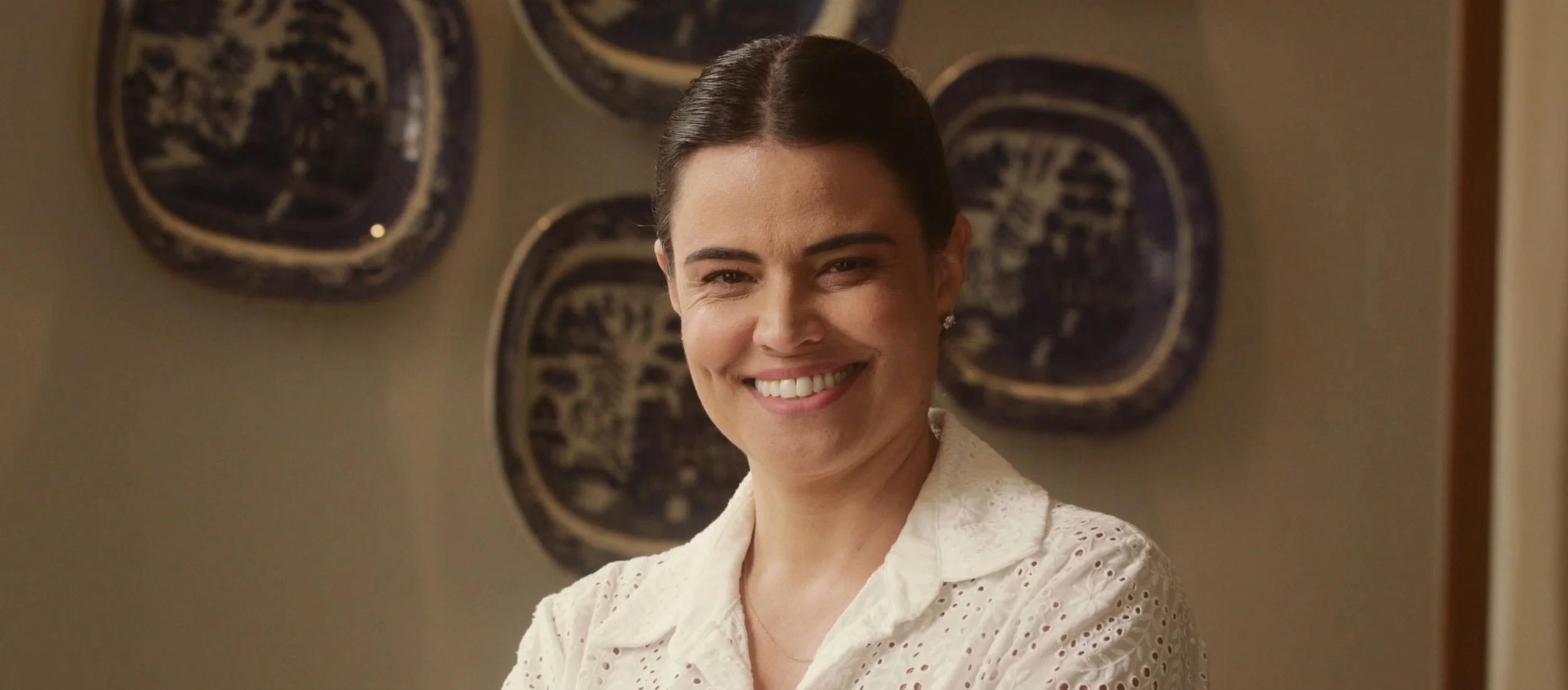 A woman with dark hair styled in a 1950s fashion, smiling and wearing a white eyelet blouse, standing in front of a wall decorated with dark blue ceramic plates.