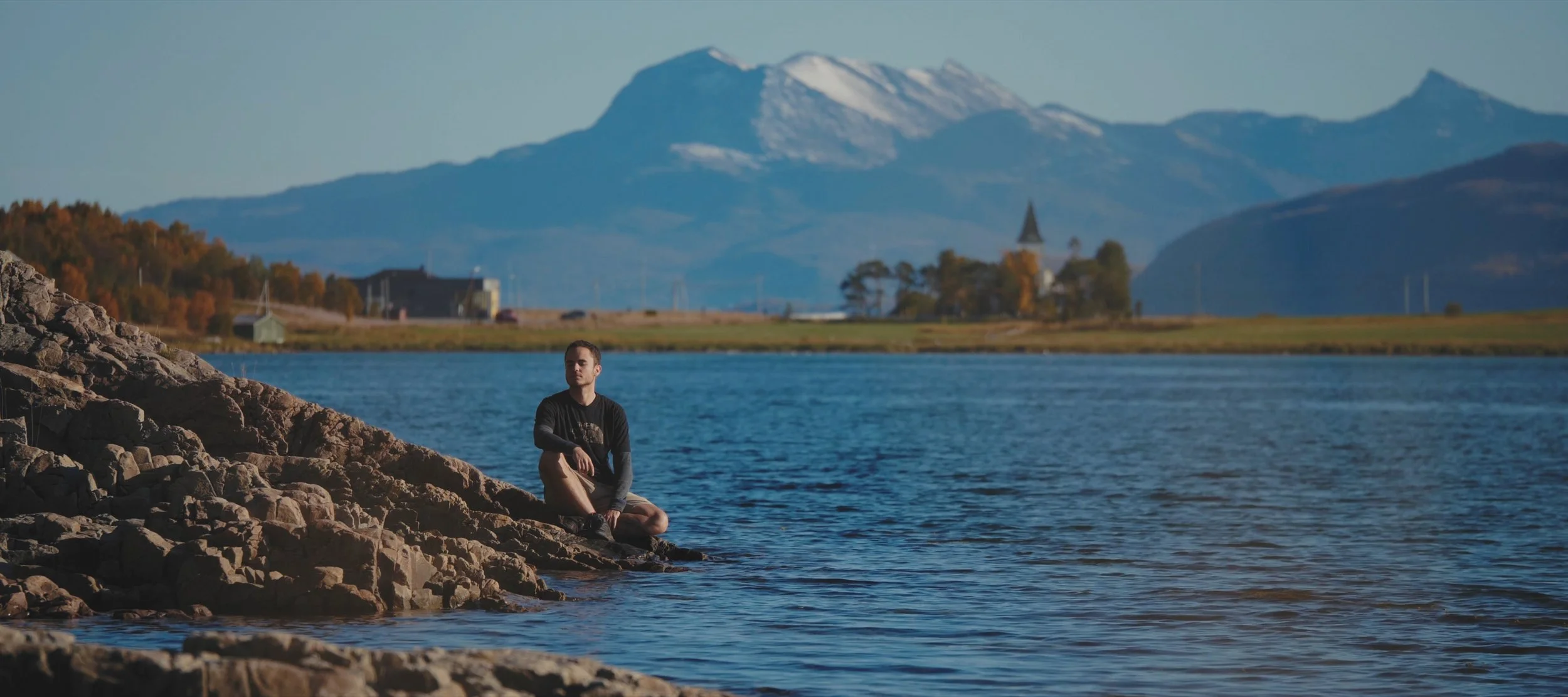 A person sitting on rocks at the edge of a lake with mountains in the background.