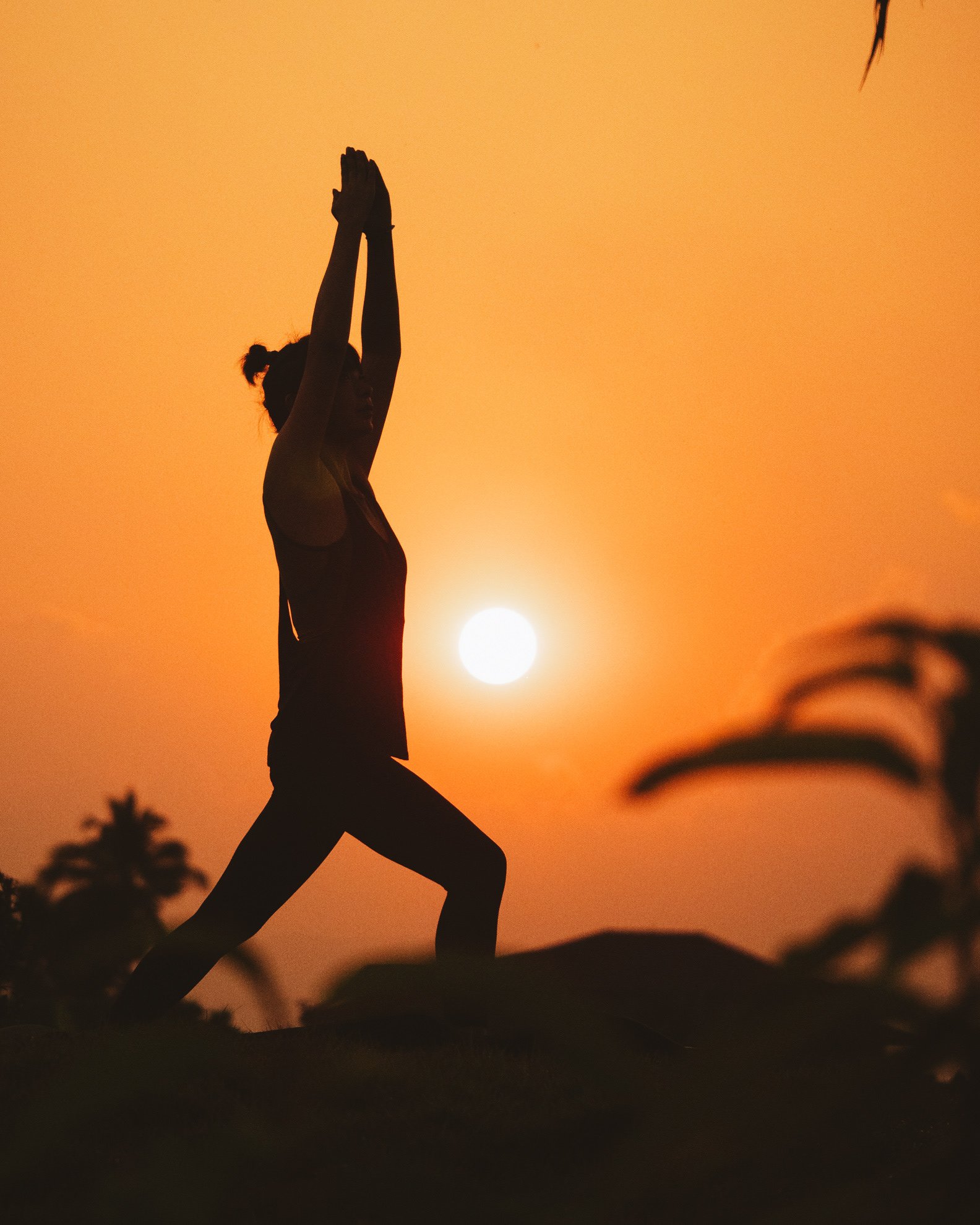 A person practicing yoga outdoors at sunset, performing a warrior pose with hands raised above head against a bright orange sky.