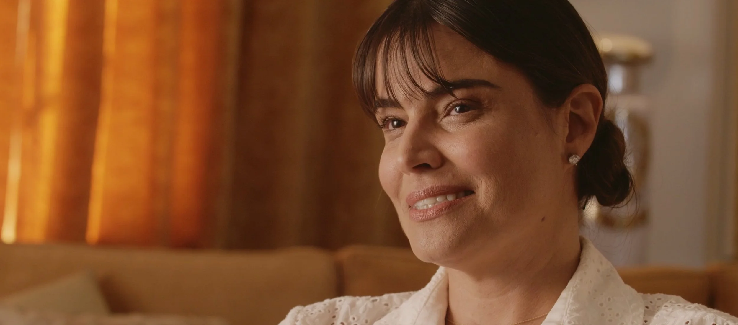 Close-up of a middle-aged woman with dark hair and earrings, smiling softly indoors with warm lighting and curtains in the background.