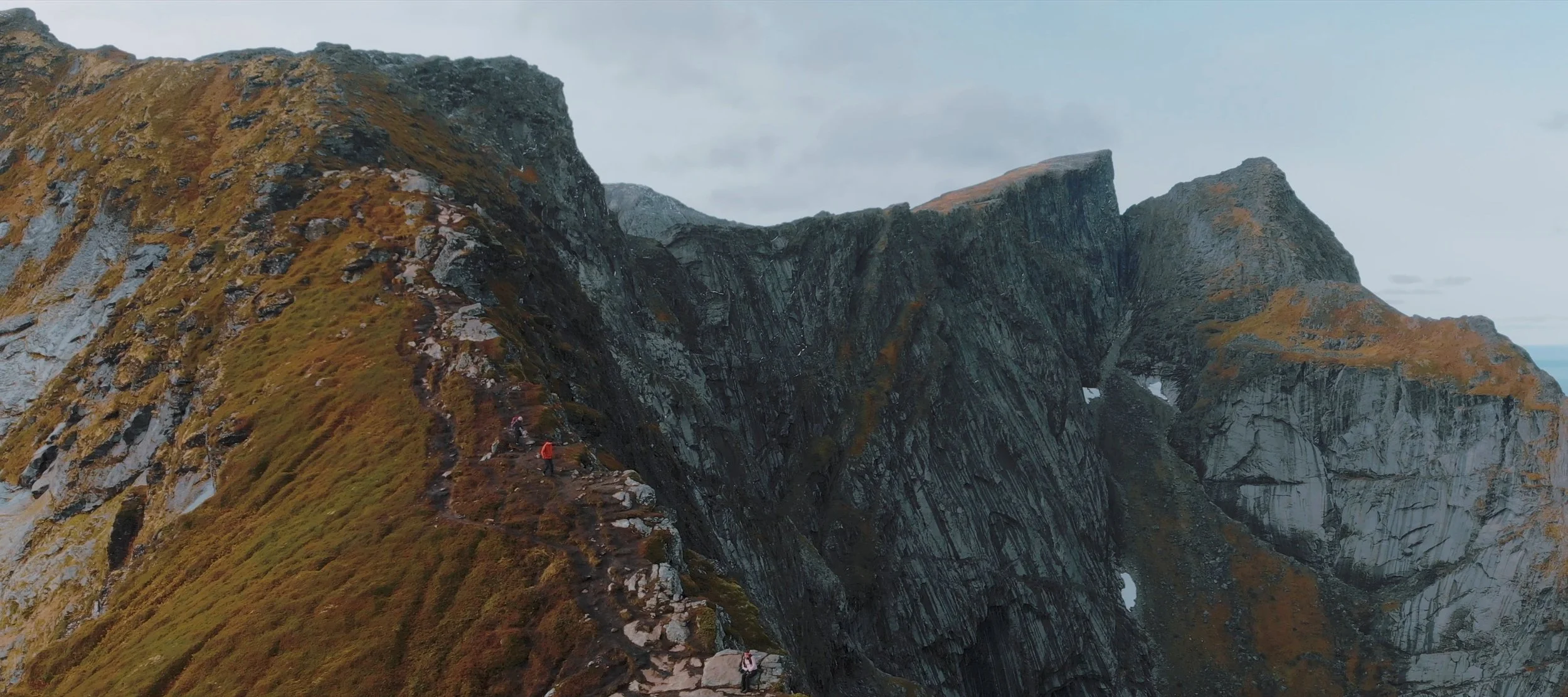 Hikers walking along a narrow mountain trail on a steep, rugged ridge with rocky cliffs and patches of grass and moss.