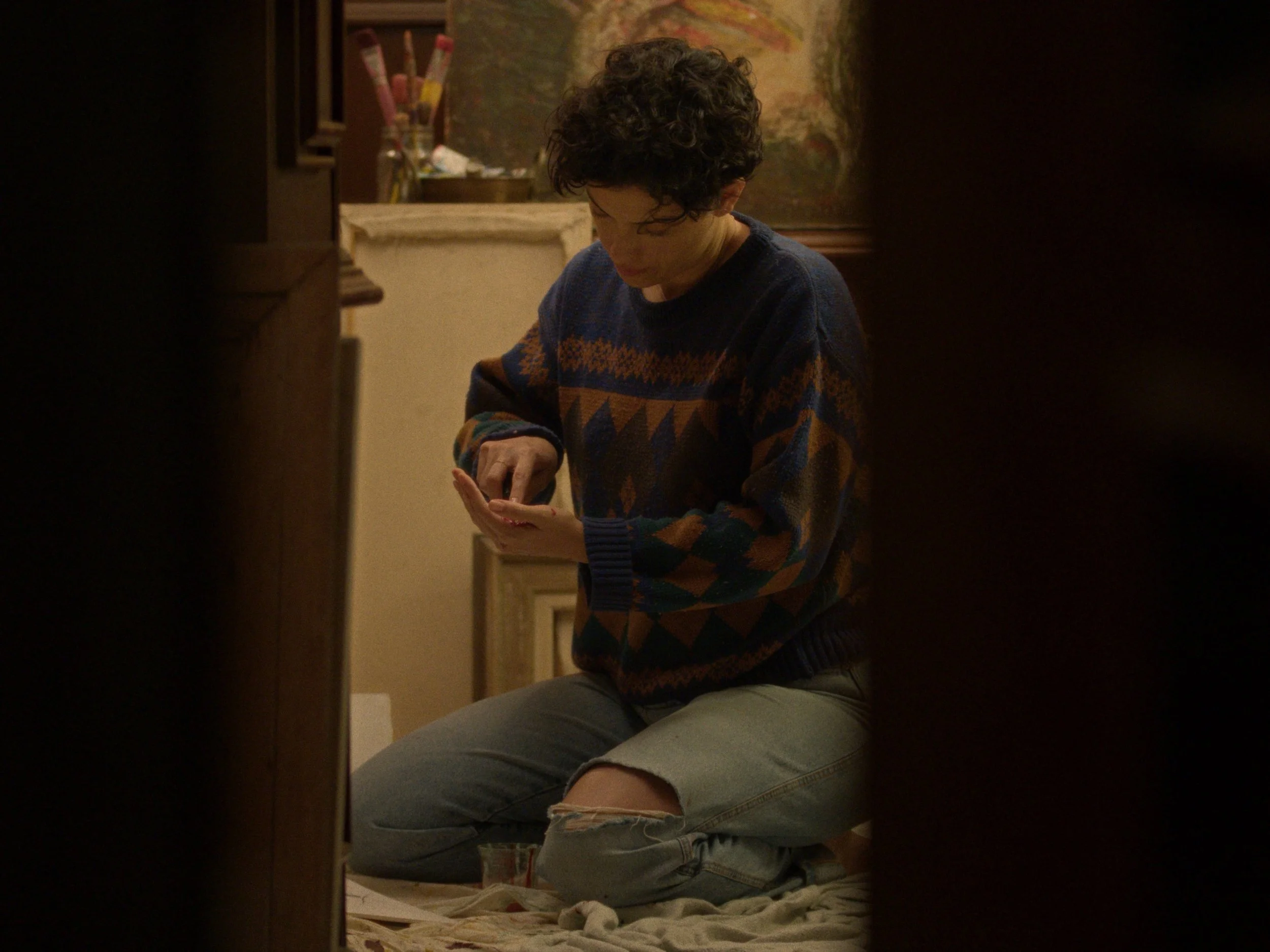 A person with curly hair, wearing a patterned sweater and ripped jeans, kneeling on the floor and looking at their hand in a room with art supplies and paintings.