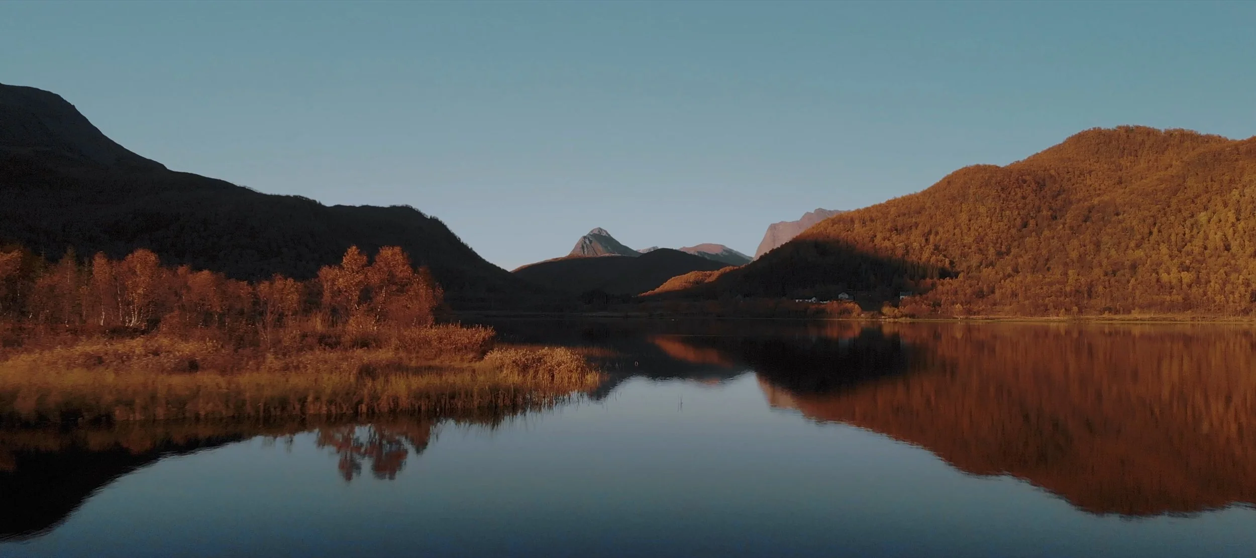 A tranquil lake reflecting orange and green autumn trees on the shoreline, with mountains in the background under a clear blue sky.