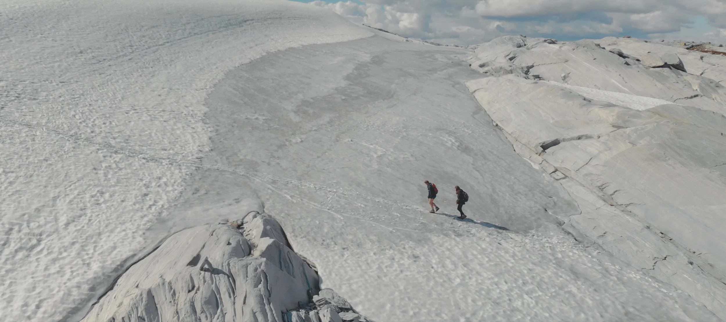 Two hikers walking on a snow and ice-covered mountain with rocks in the background.