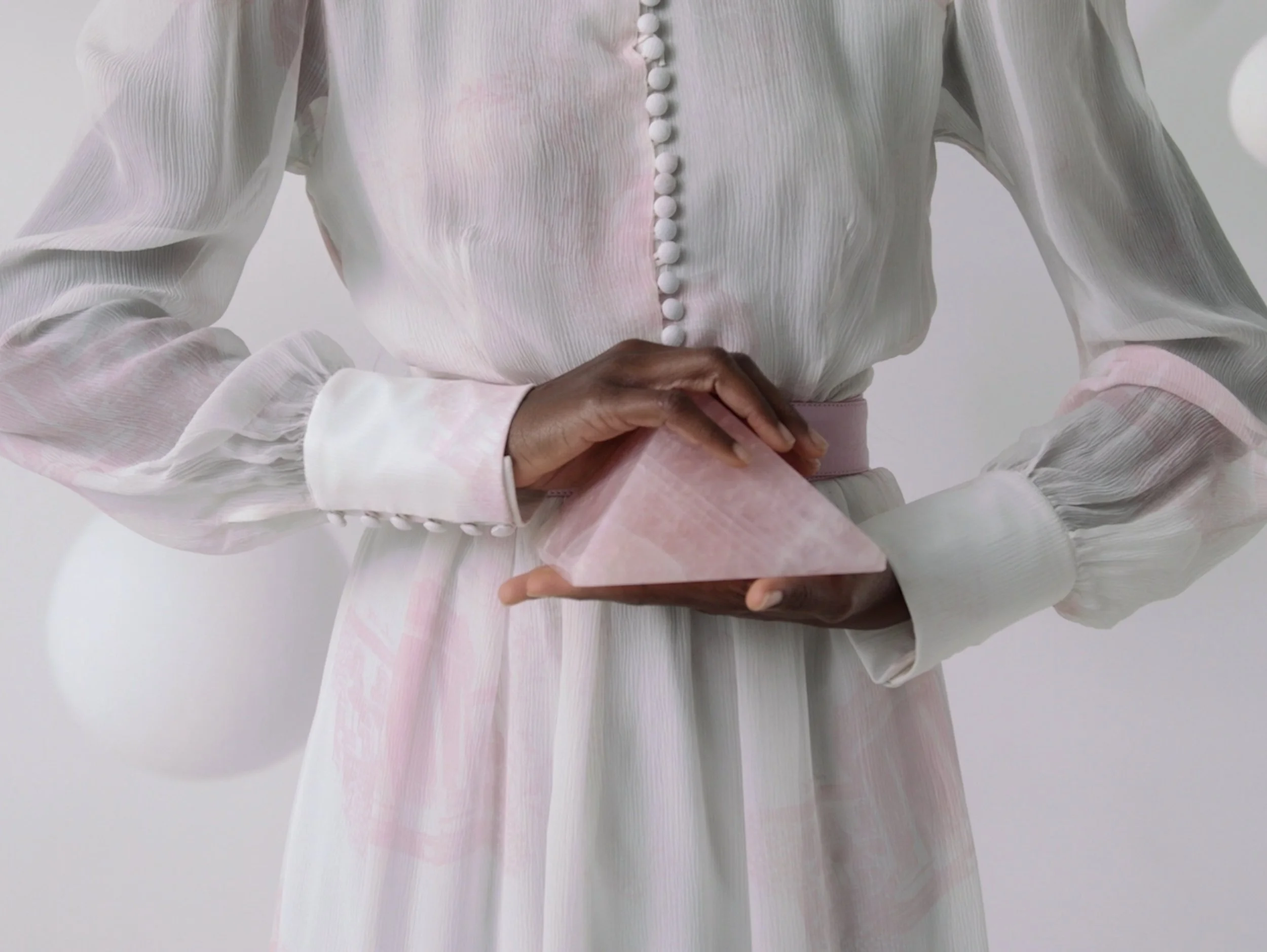 A woman in a light pink and white dress is holding a pink marble pyramid sculpture.