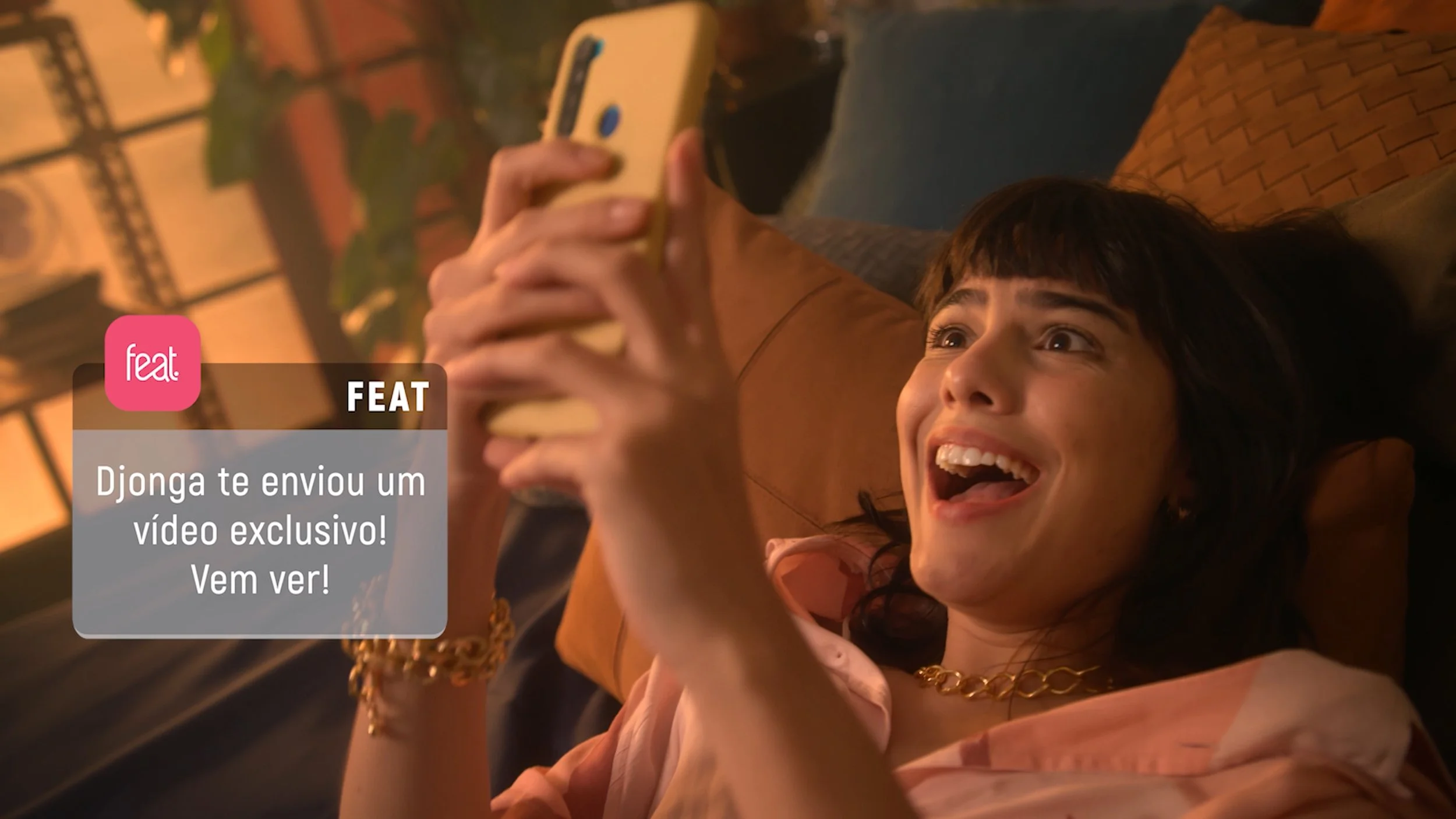 A woman with dark hair and a gold chain necklace, lying on a couch and taking a selfie with a smartphone, smiling with her mouth open. There is a message overlay in Portuguese that reads, "Djonga te envia um vídeo exclusivo! Vem ver!".