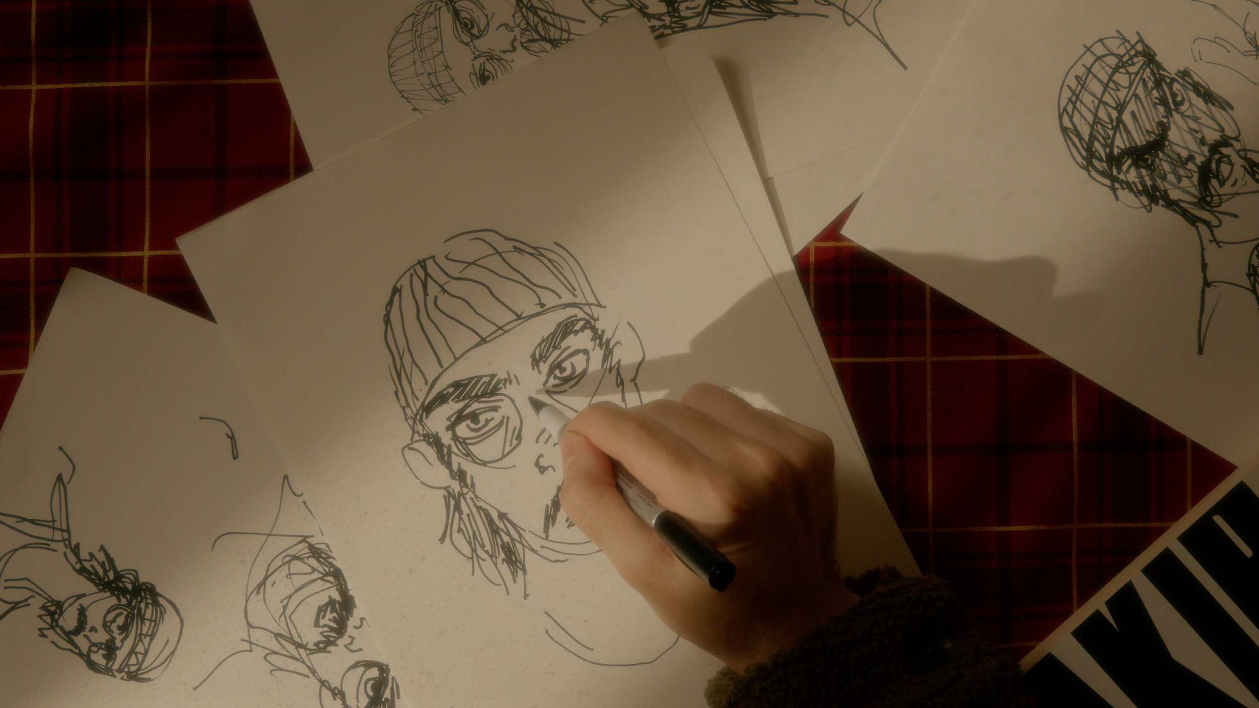 Person sketching a portrait of a man with glasses and a beanie on white paper, surrounded by other sketches of faces, on a table with a red plaid tablecloth.