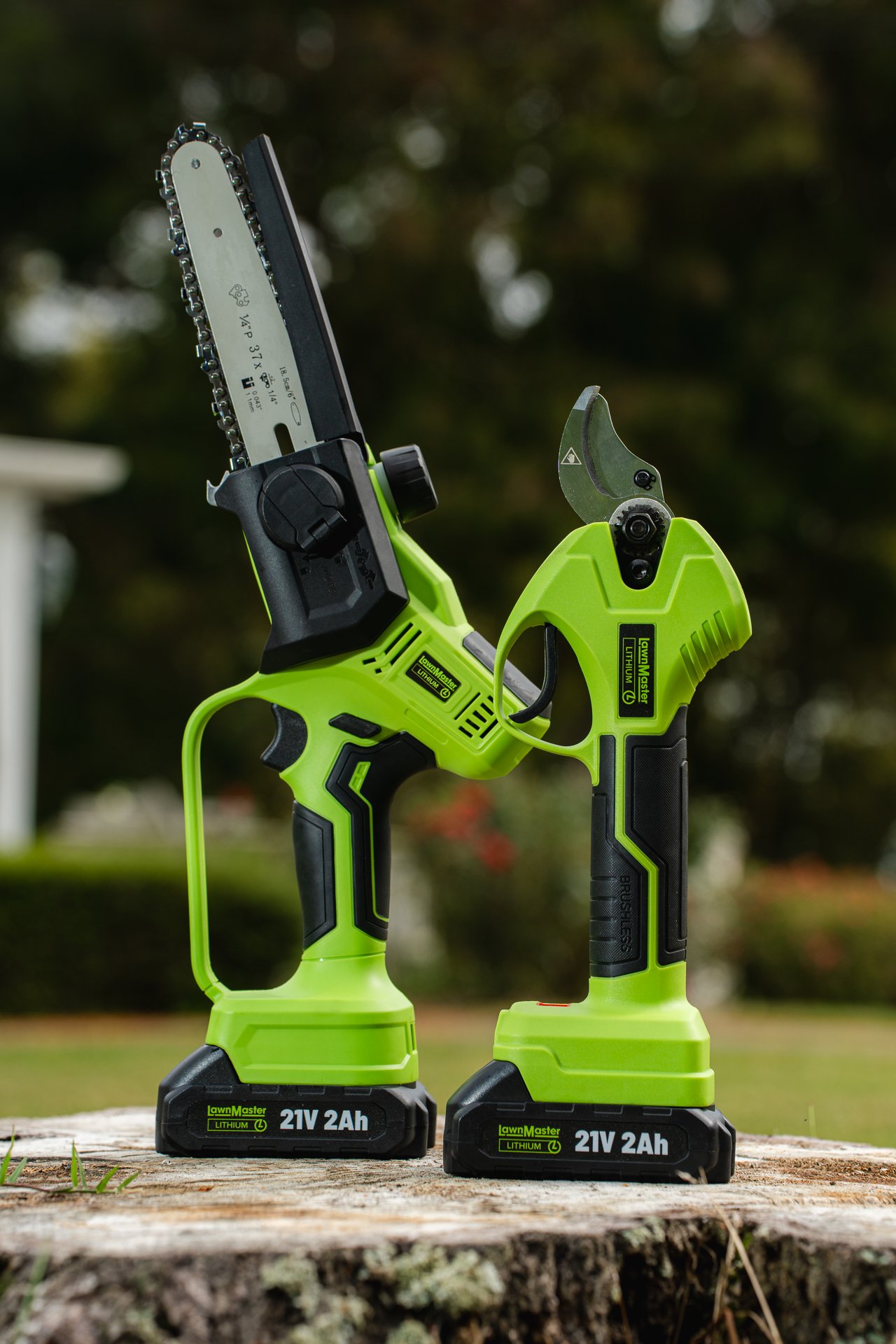Green cordless electric pruning shears and hedge trimmers placed on a tree stump outdoors with blurred trees and sky in the background.