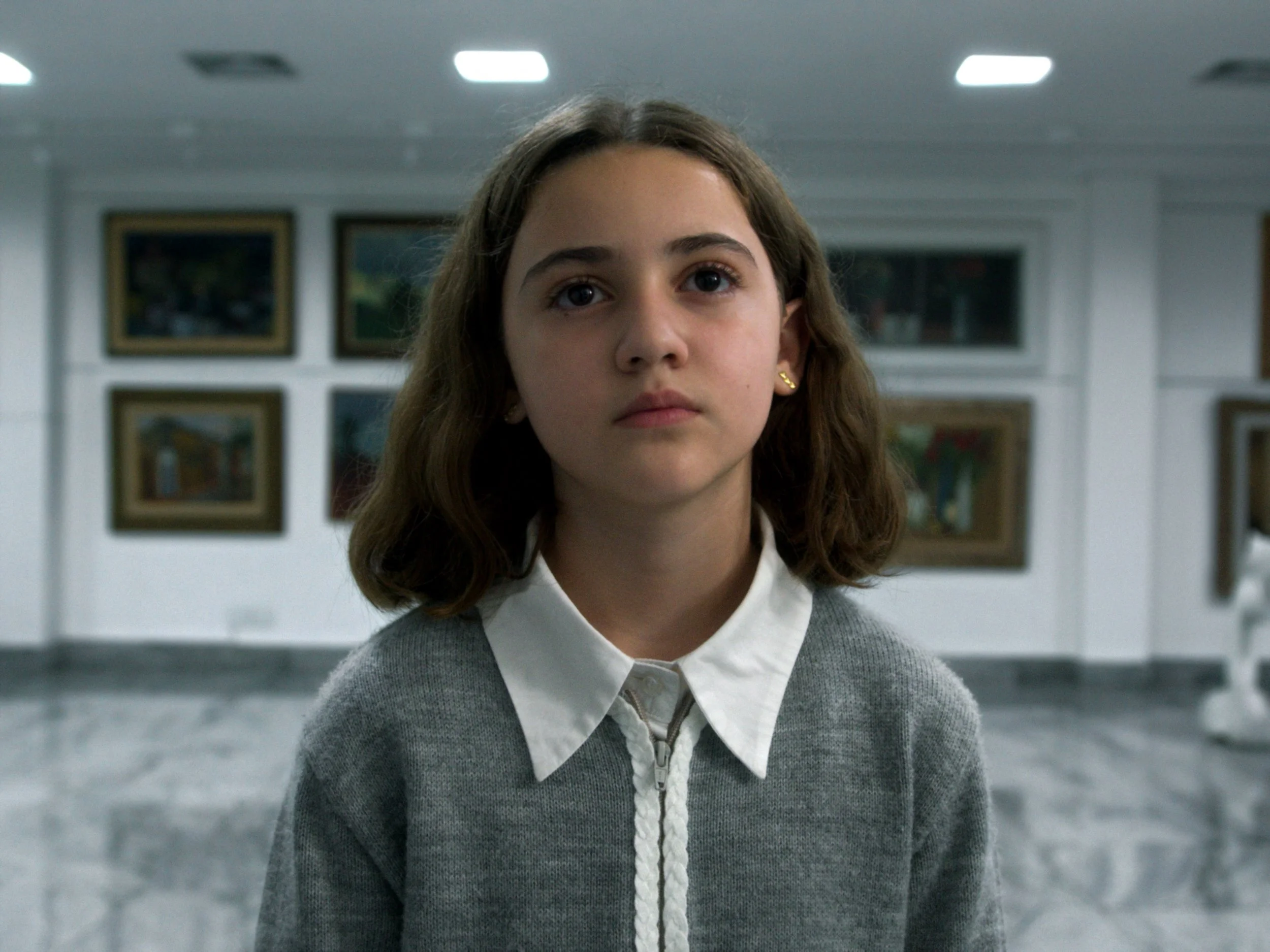 A young girl with brown hair, wearing a grey sweater over a white collared shirt, standing in an art gallery or museum with paintings on the wall behind her.