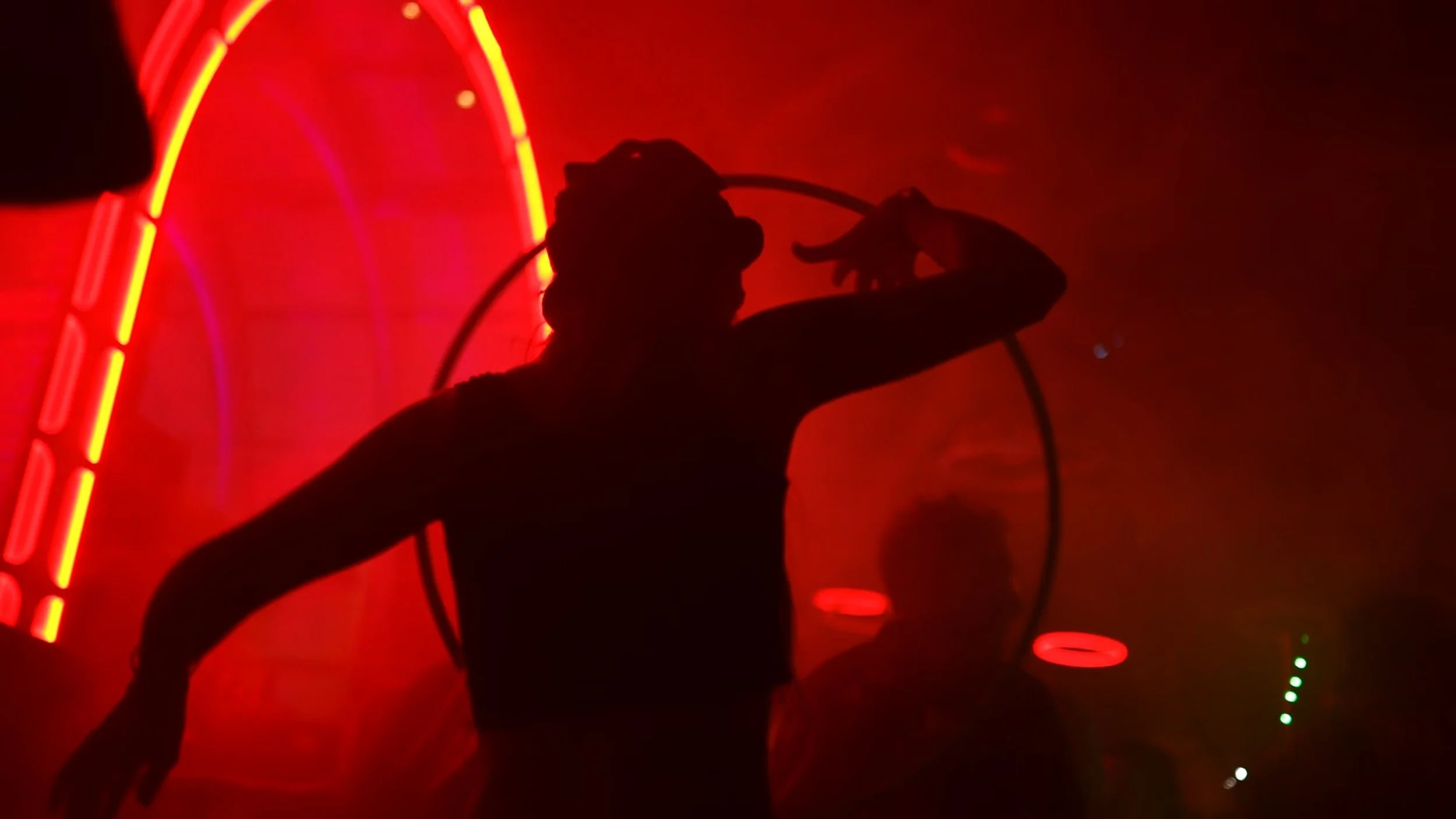 Silhouette of a person in a dark room with red neon lighting, posing with one arm extended and fingers pointing, with a large hoop around their head.