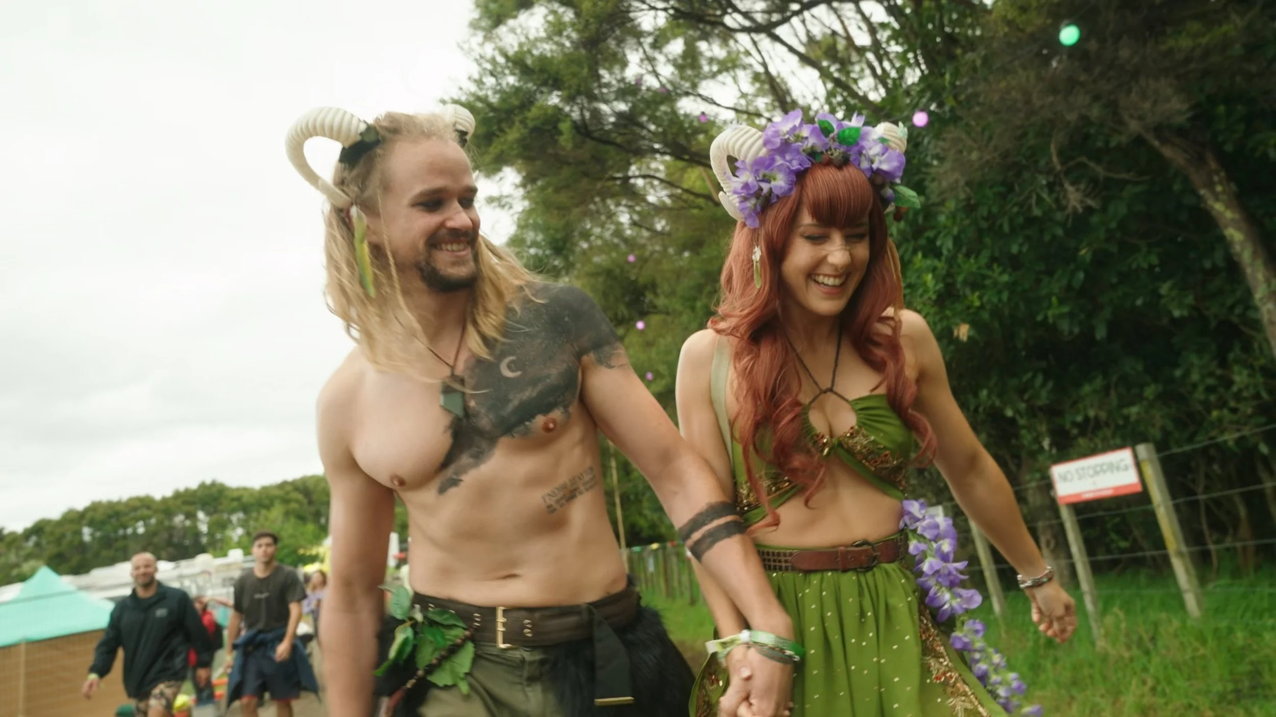 Two people dressed as mythical forest characters, one with ram horns and tattoos, the other with a flower crown, holding hands and smiling outdoors.