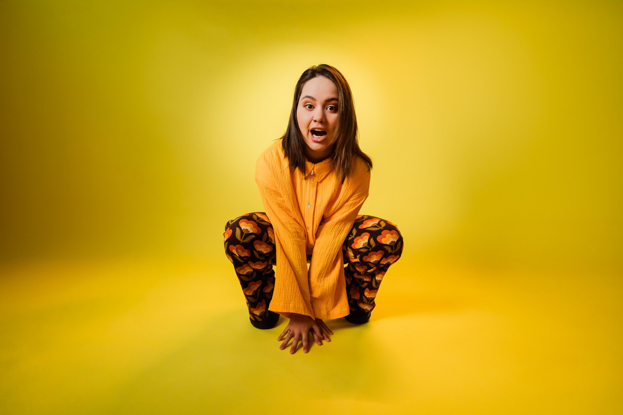 Young woman crouching on a yellow background, wearing an orange shirt and patterned pants, with an expressive surprised facial expression.