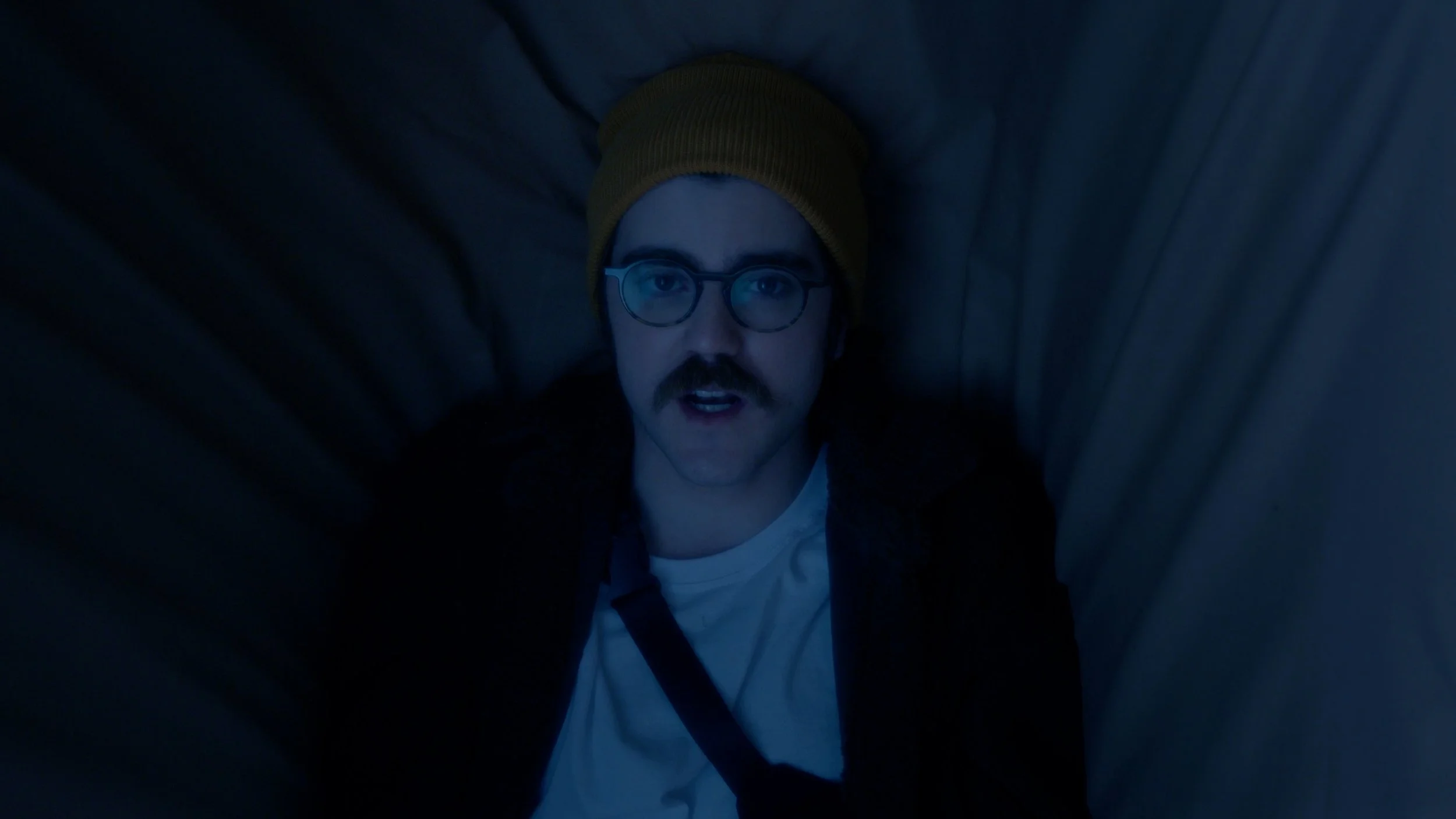 A man with glasses, a mustache, and a beard looks up into the camera while lying down in a narrow, dark space, wearing a yellow beanie and a white shirt.