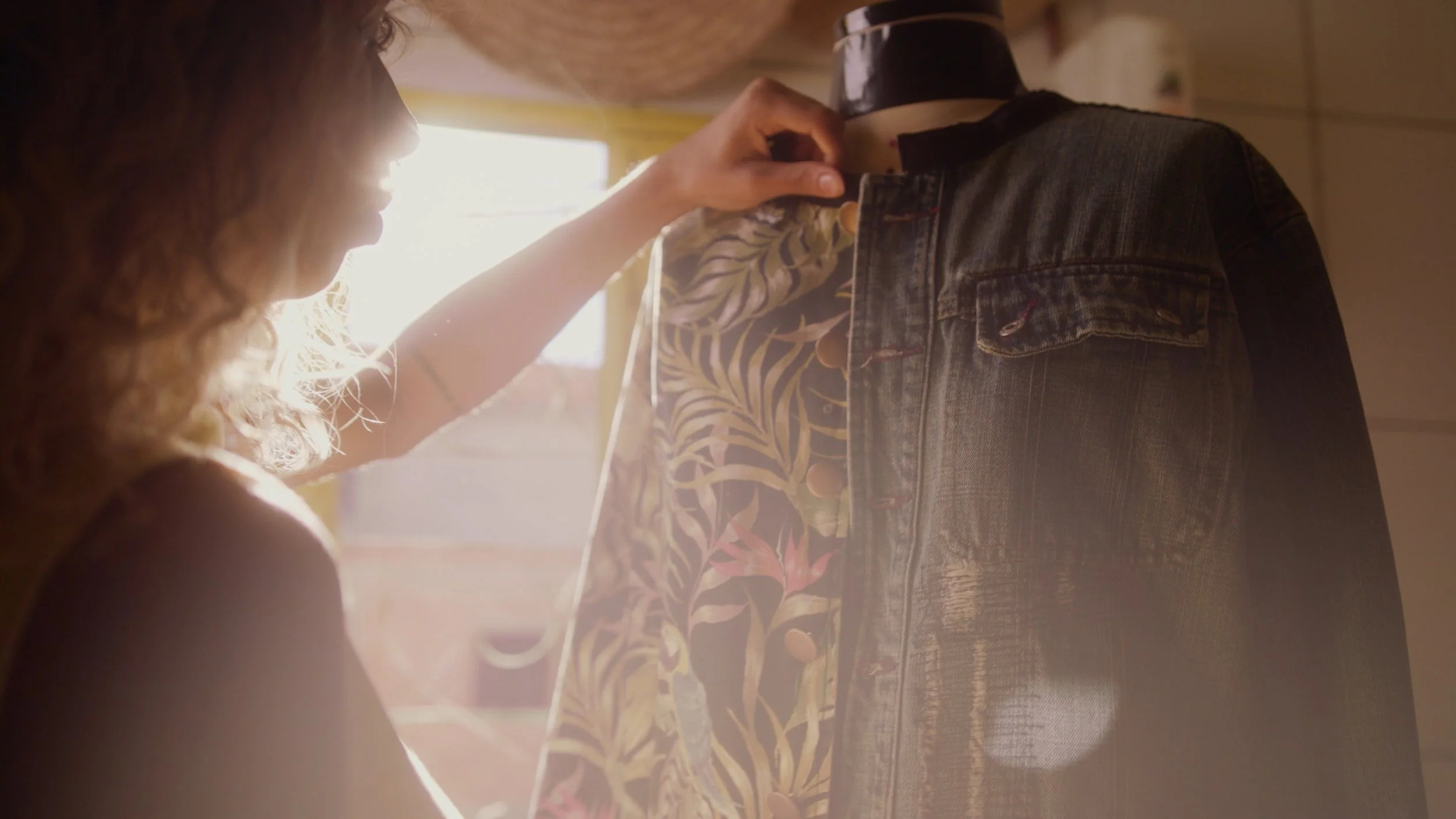 A woman adjusting a floral blouse on a mannequin wearing a denim jacket, with sunlight streaming in.