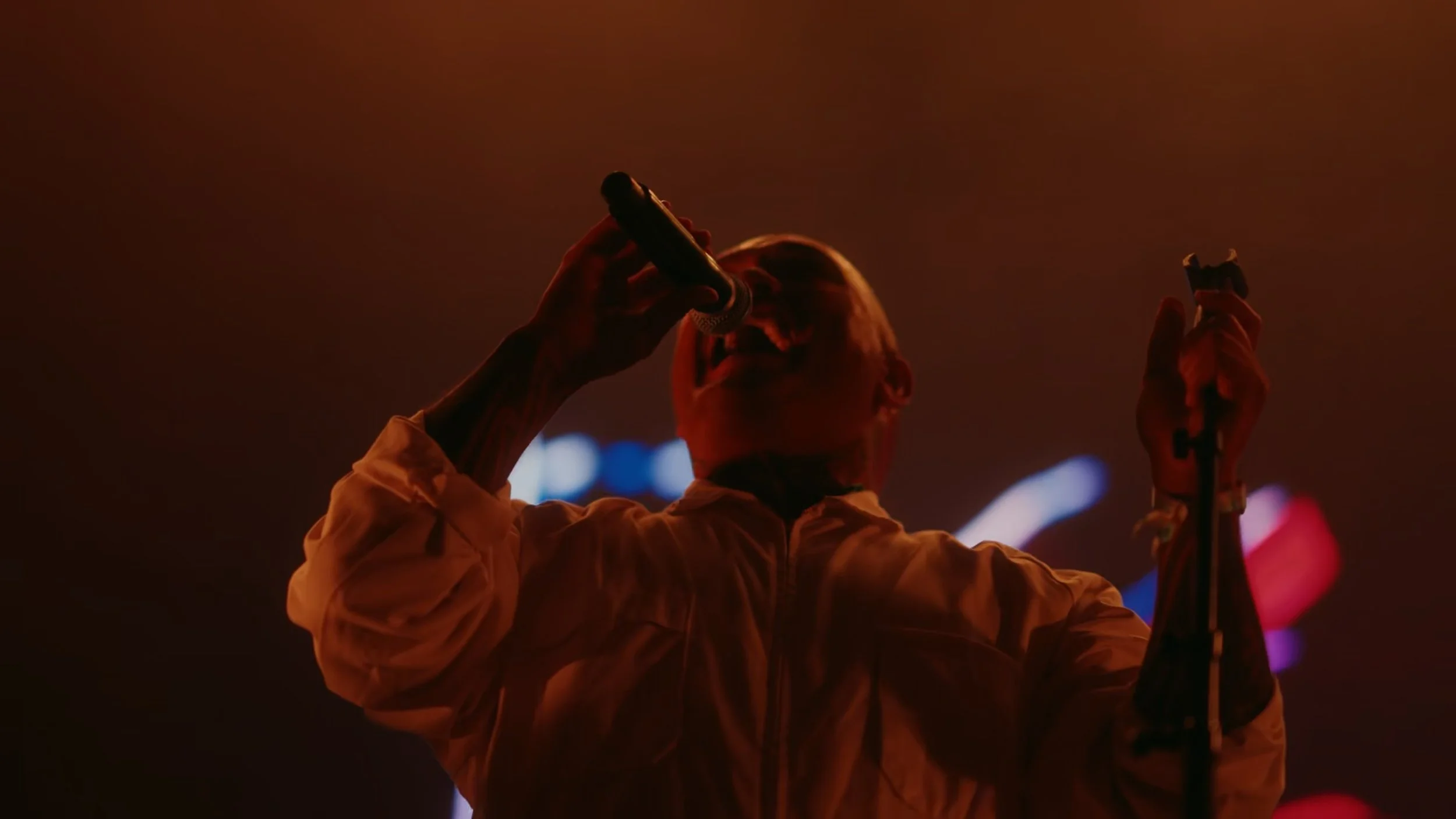 A singer performing on stage, holding a microphone and wearing a white jacket, illuminated by red stage lighting.