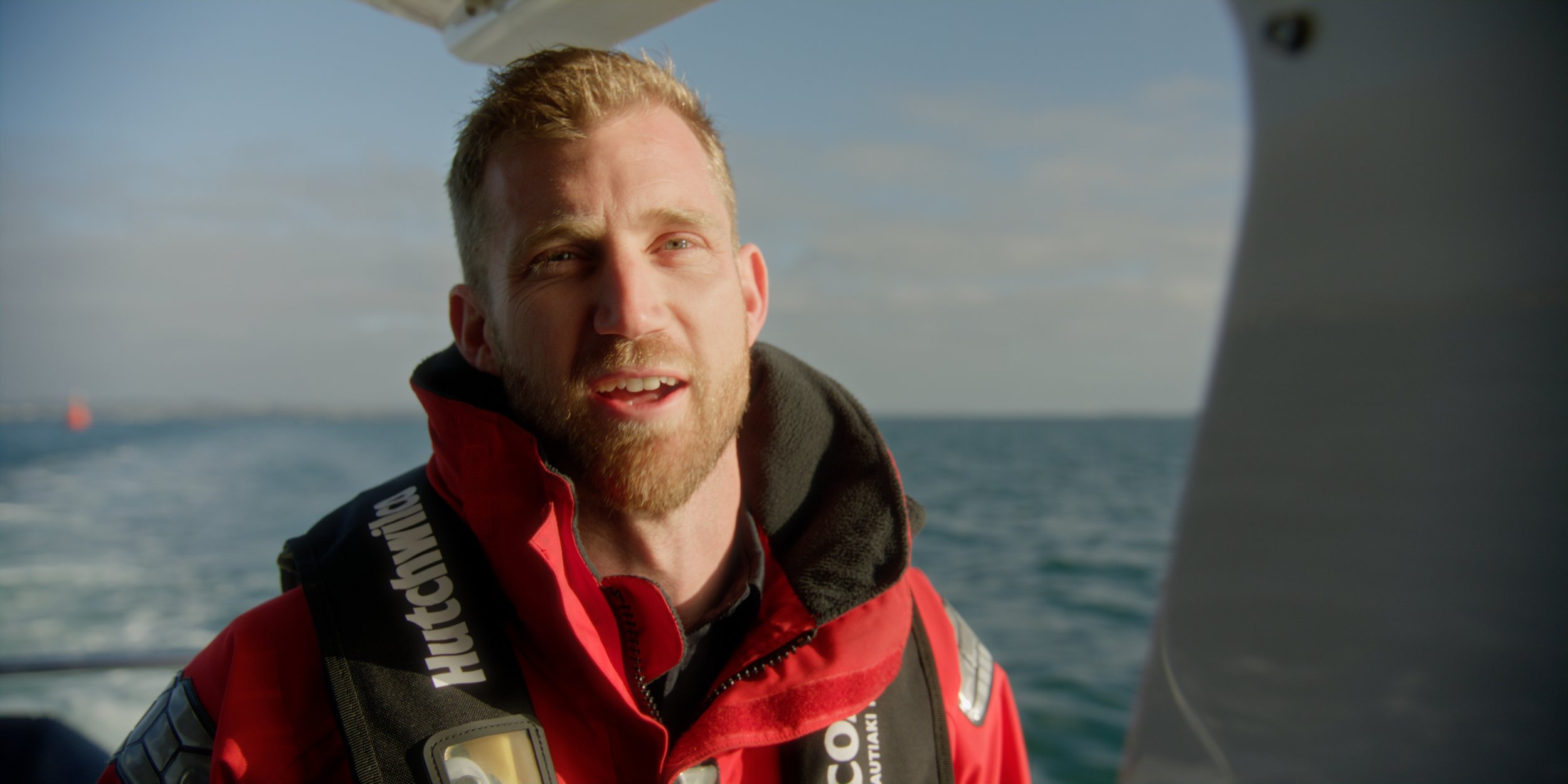 A man with blonde hair and a beard in a red and black jacket on a boat, with water and a cloudy sky in the background.
