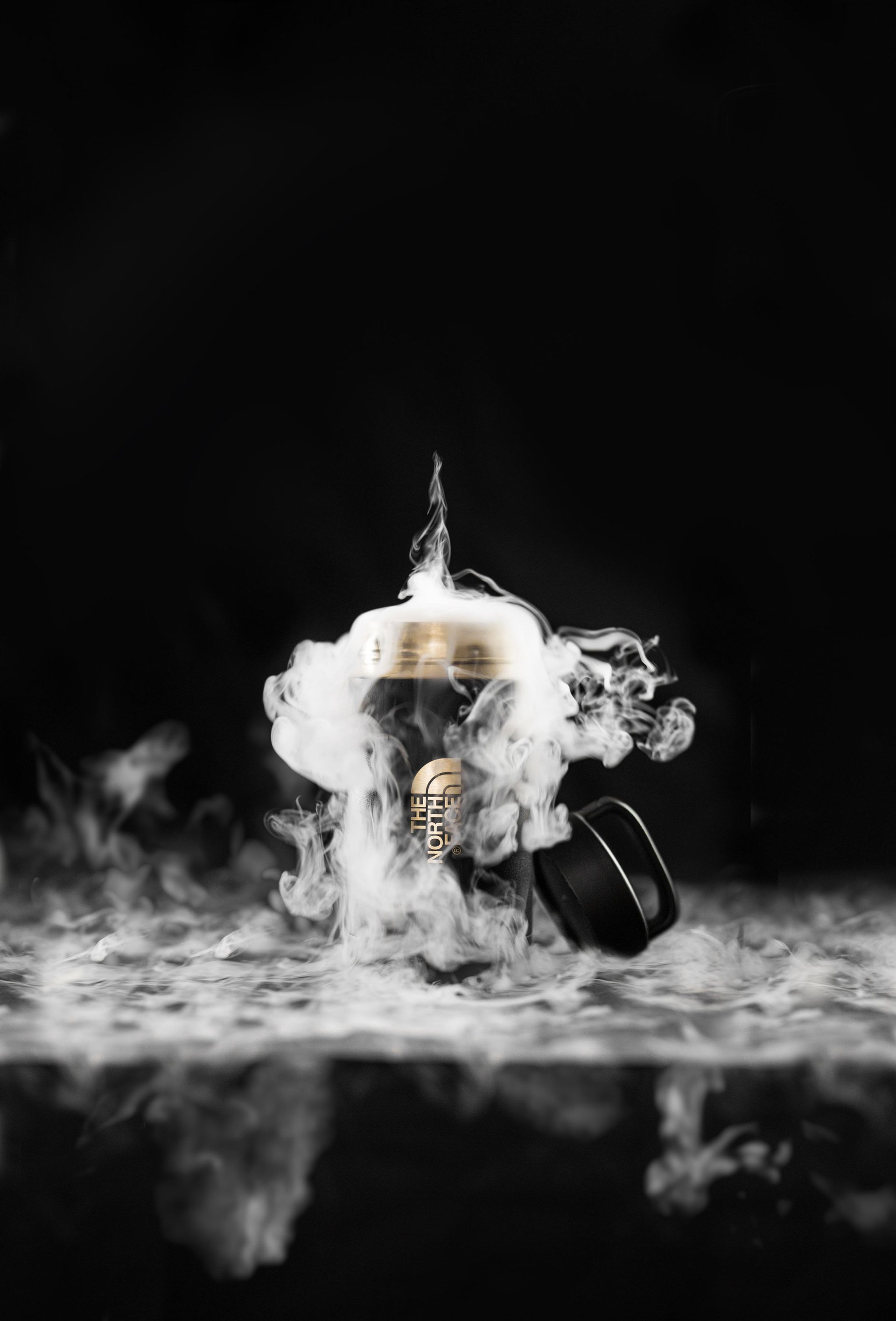 A camera with the North Face logo surrounded by swirling smoke, set on a reflective surface against a black background.